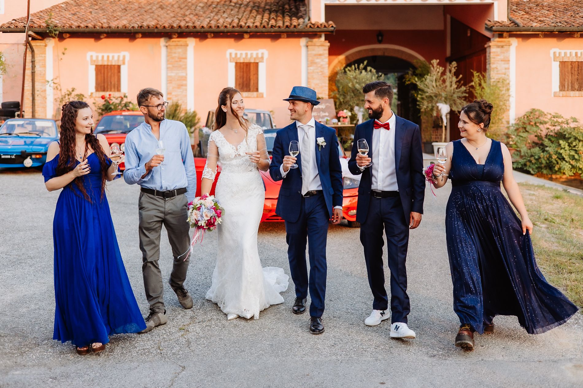 A wedding party walks together laughing and smiling outside a rustic Italian villa with blue and white formal attire.
