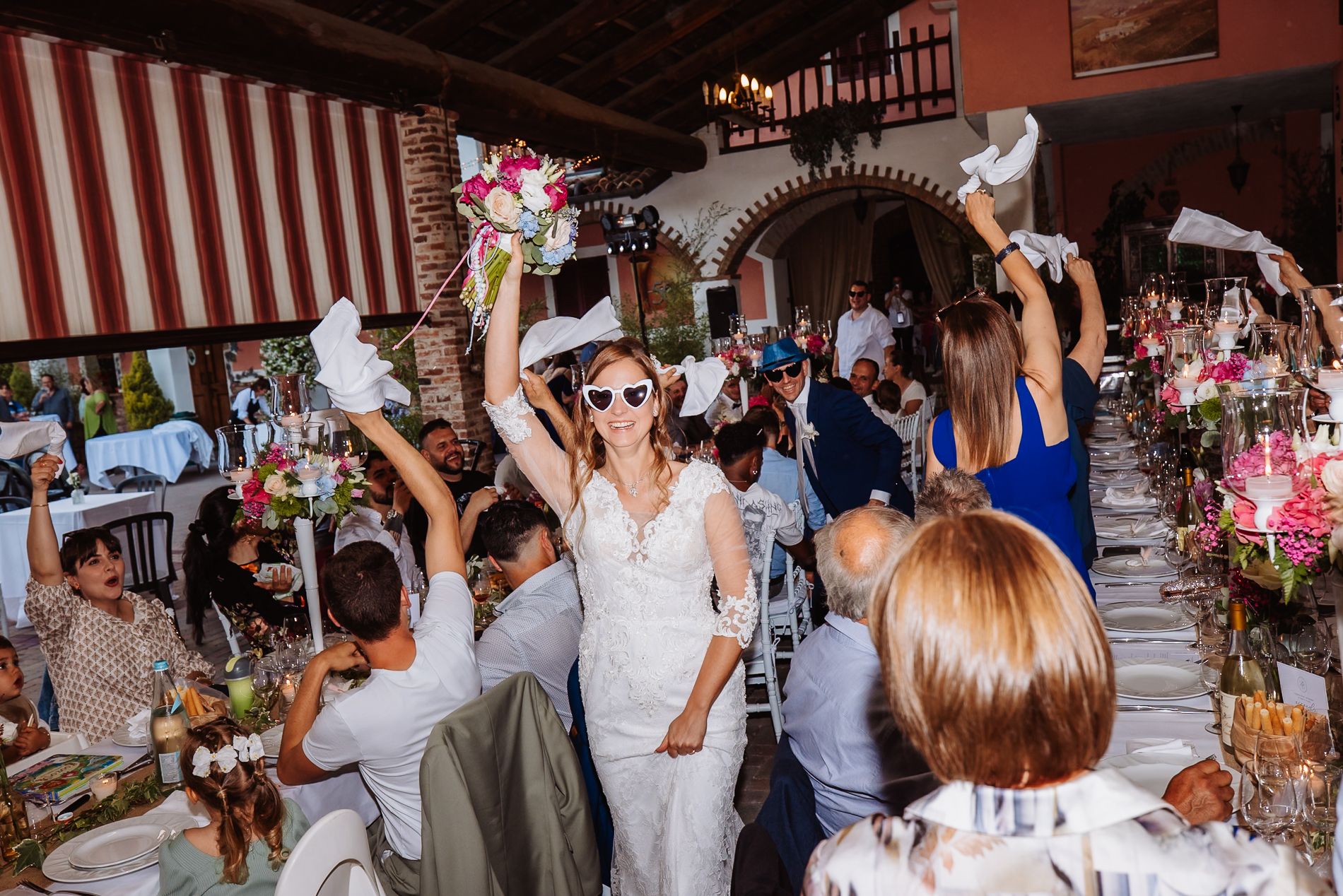 Wedding celebration with guests waving white napkins during reception in rustic venue with striped awning.