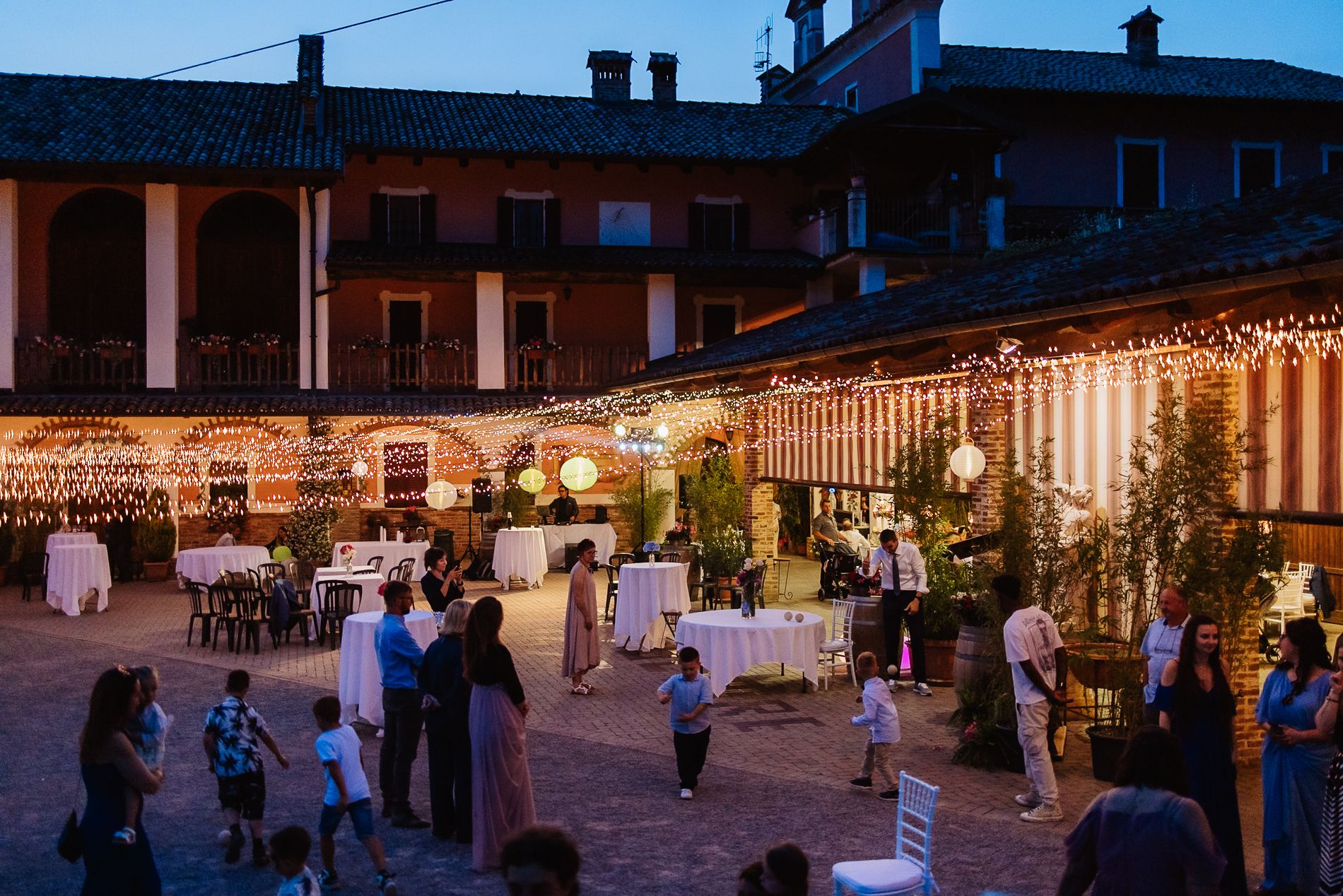 An outdoor evening wedding reception with string lights decorating a terrace area where guests mingle around tables.