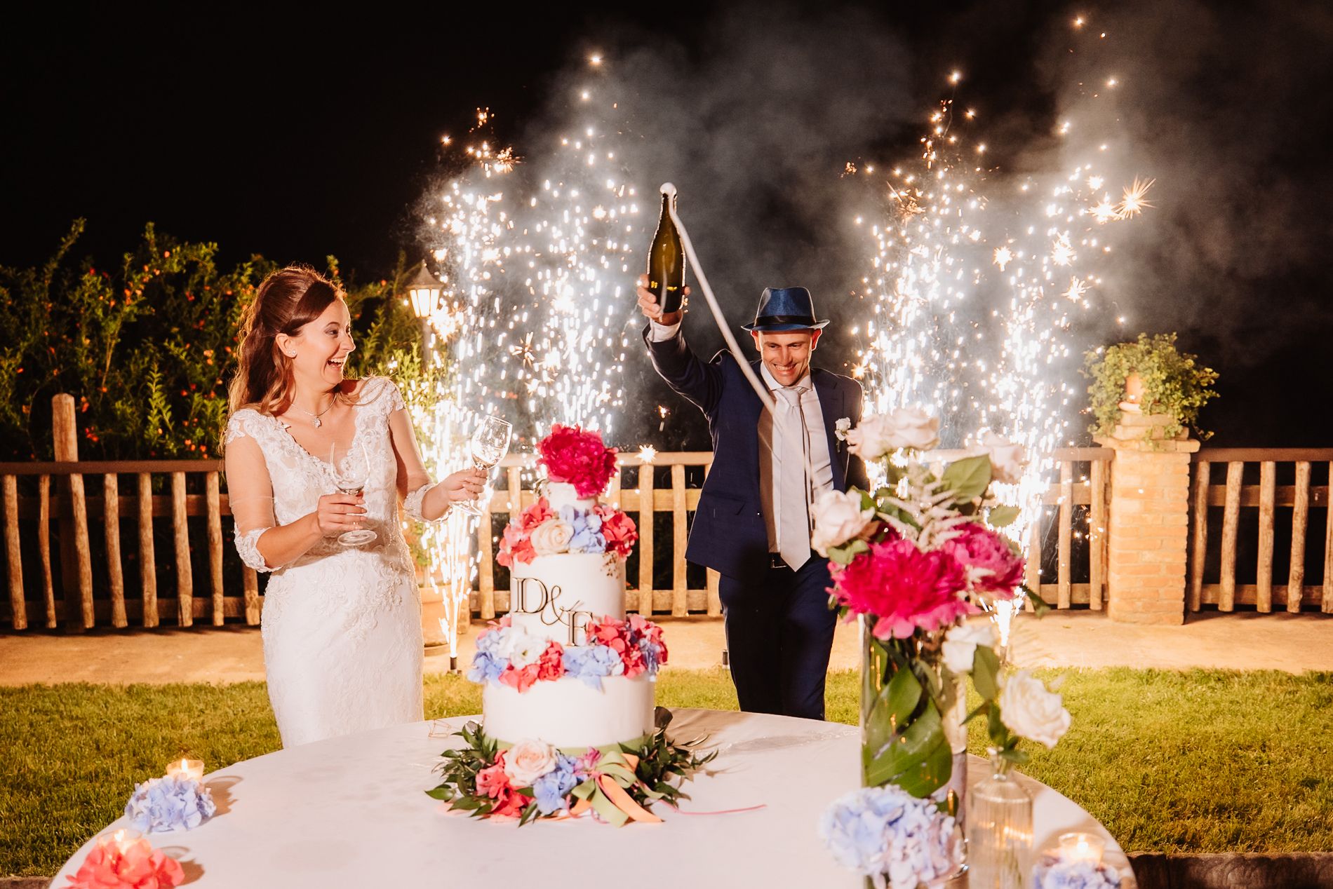 Wedding celebration at night with sparklers illuminating a multi-tiered cake decorated with fresh flowers on an outdoor table.