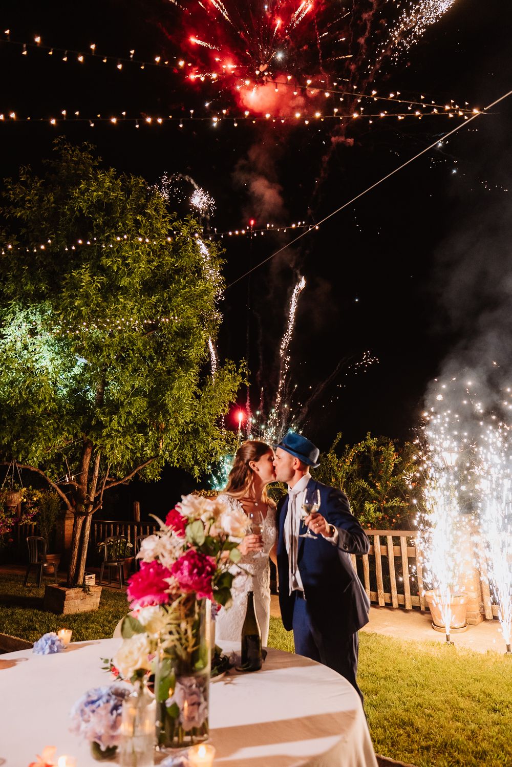 A couple celebrates at an outdoor wedding reception with fireworks lighting up the night sky while standing near floral centerpieces.