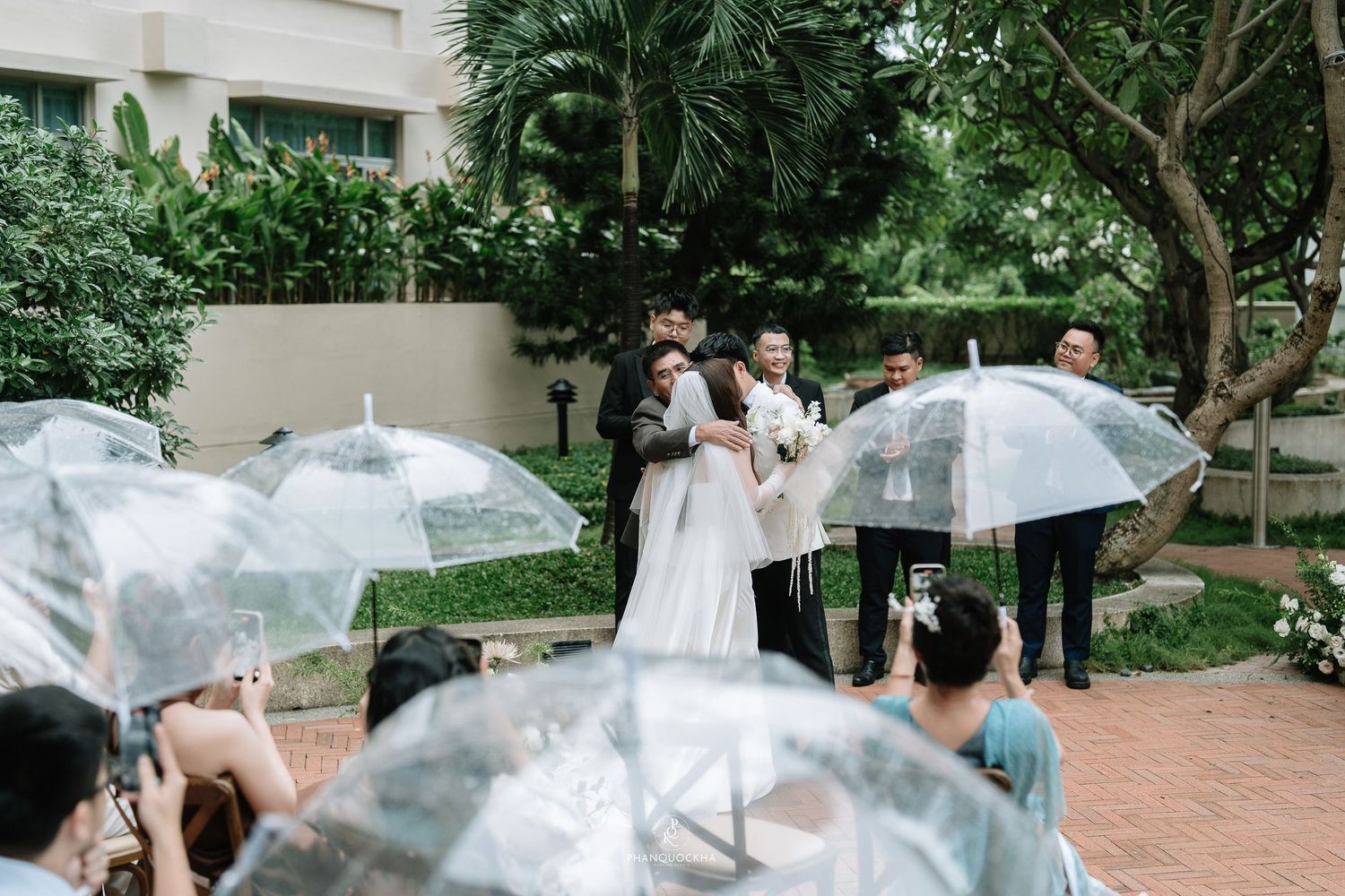 Bride and groom share embrace surrounded by guests holding clear umbrellas at outdoor rainy wedding ceremony.