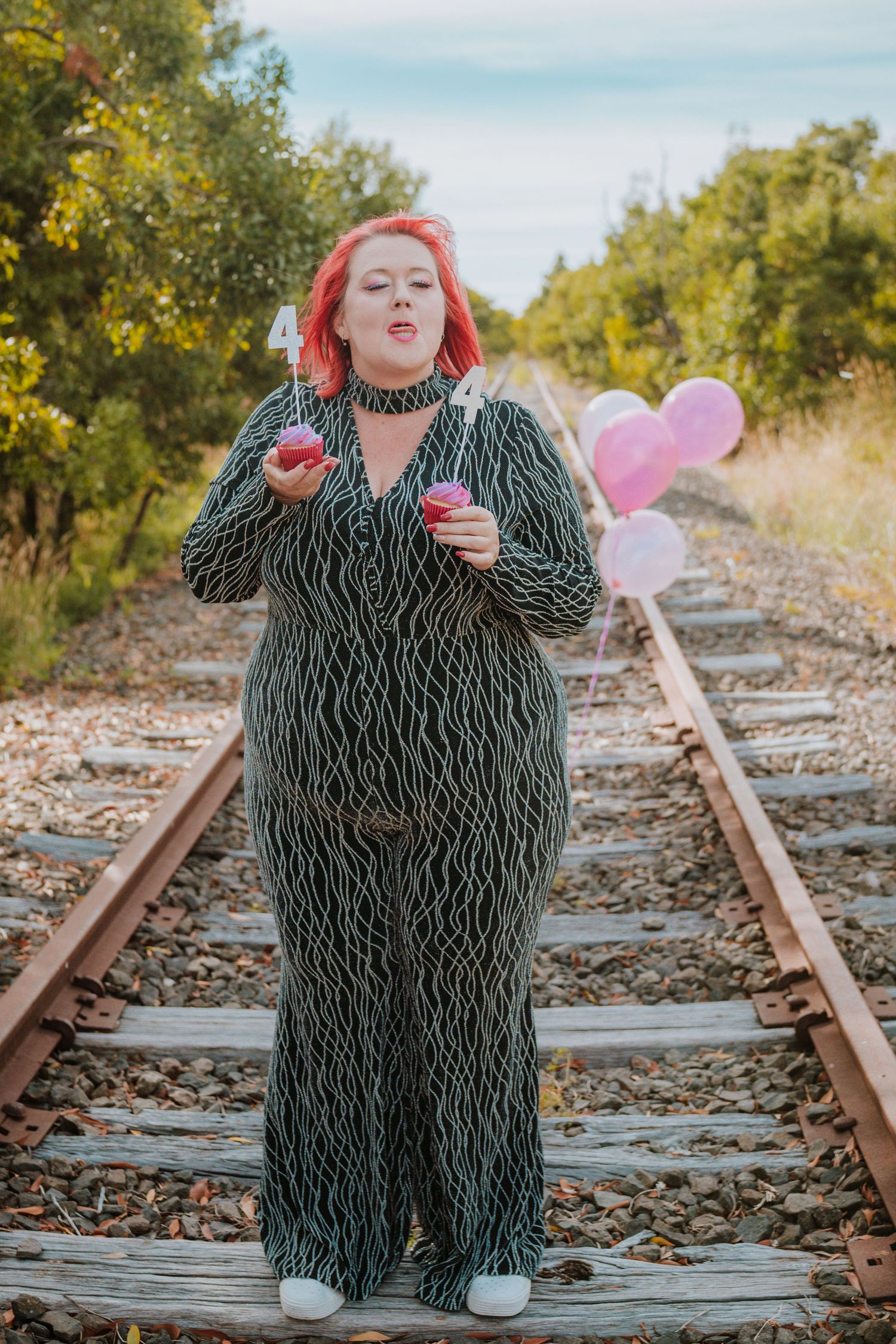 A person in a striped jumpsuit poses on railroad tracks with pink balloons in a sunny outdoor setting.