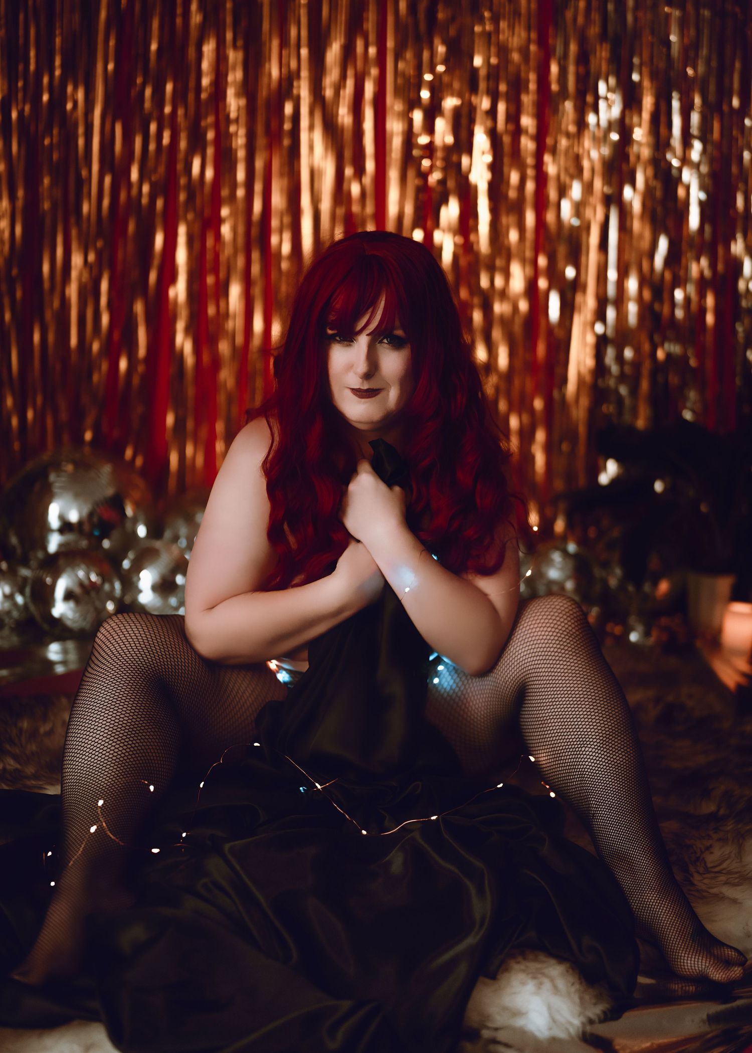 A person in black attire with vibrant red hair poses against a shimmering gold tinsel backdrop with disco balls.
