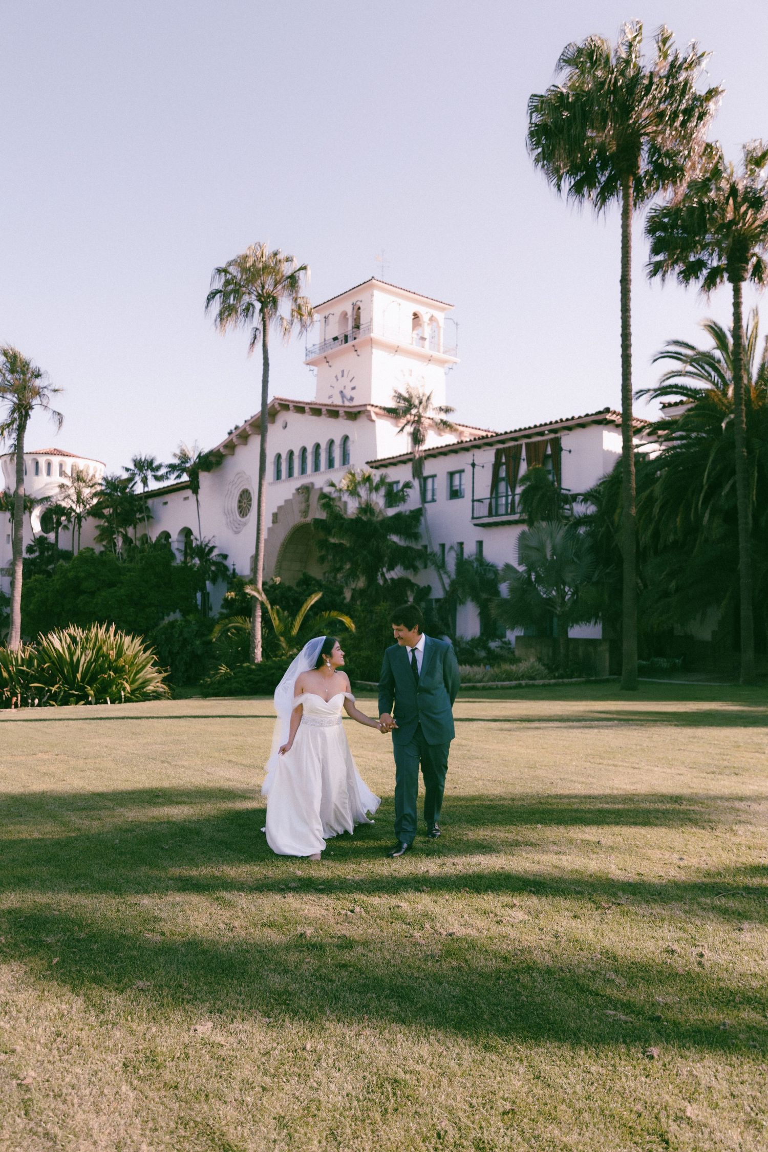 A couple in wedding attire walk on a manicured lawn in front of a Spanish-style building with palm trees and white tower.