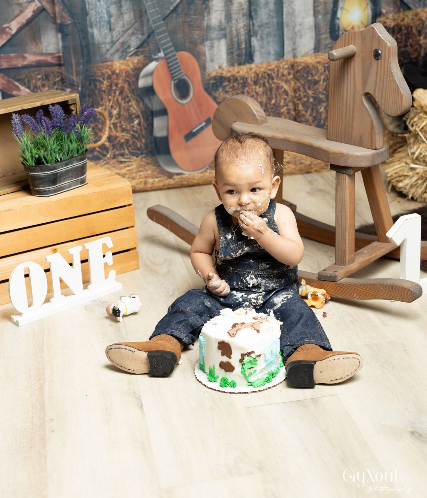 First birthday celebration setup with rustic decorations including wooden rocking horse, guitar, and smash cake against barn backdrop.
