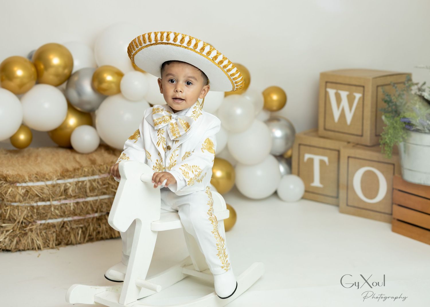 White and gold decorated outfit and hat against balloon garland backdrop with wooden letter blocks and hay bales.