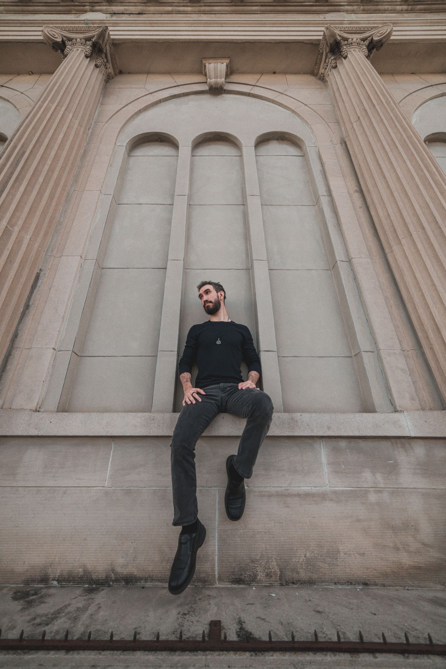 A person dressed in dark clothing sits on a stone ledge against a neoclassical architectural backdrop with columns.