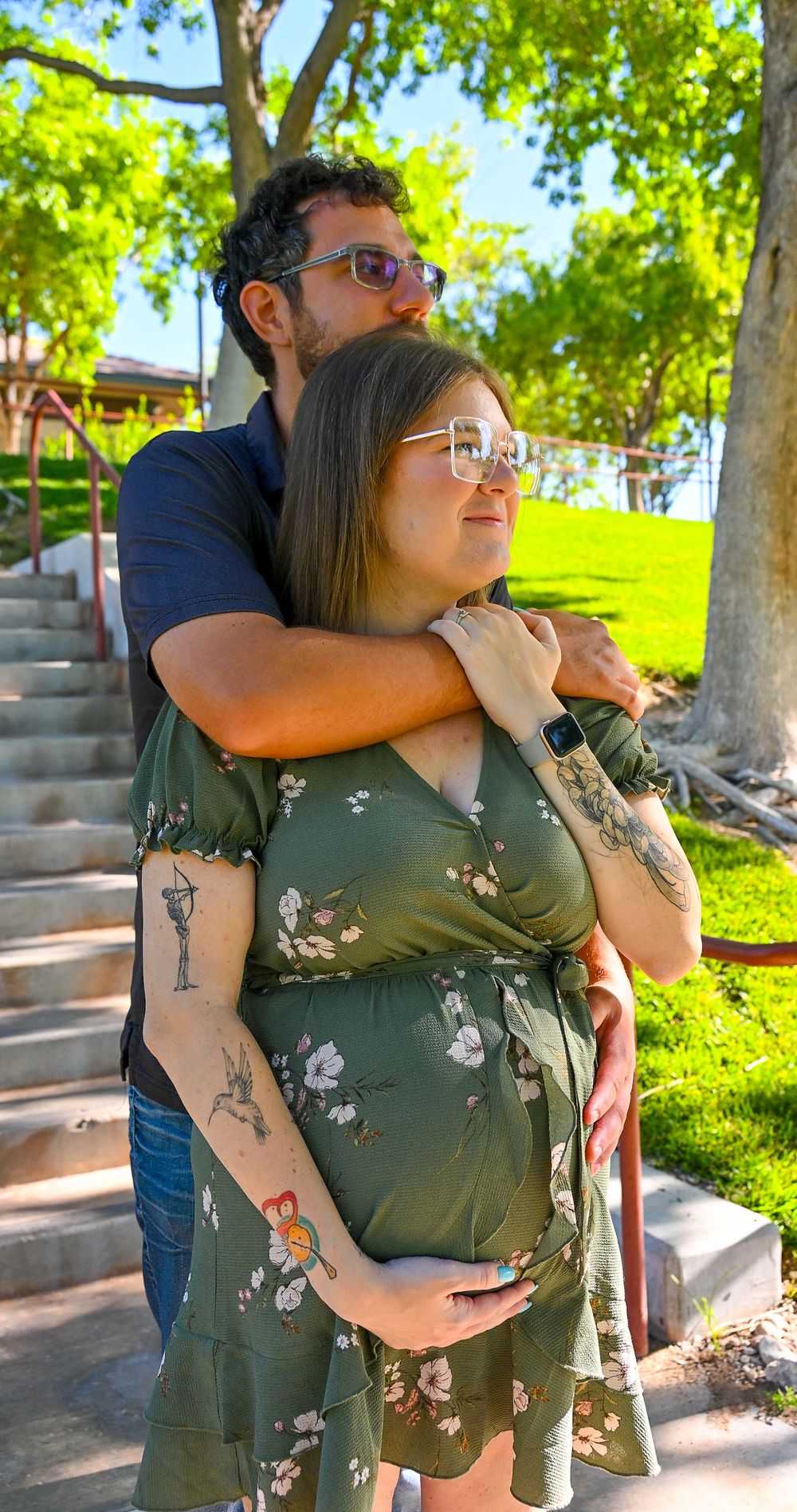 Expecting couple posing in park, man embracing pregnant woman in green floral dress from behind.