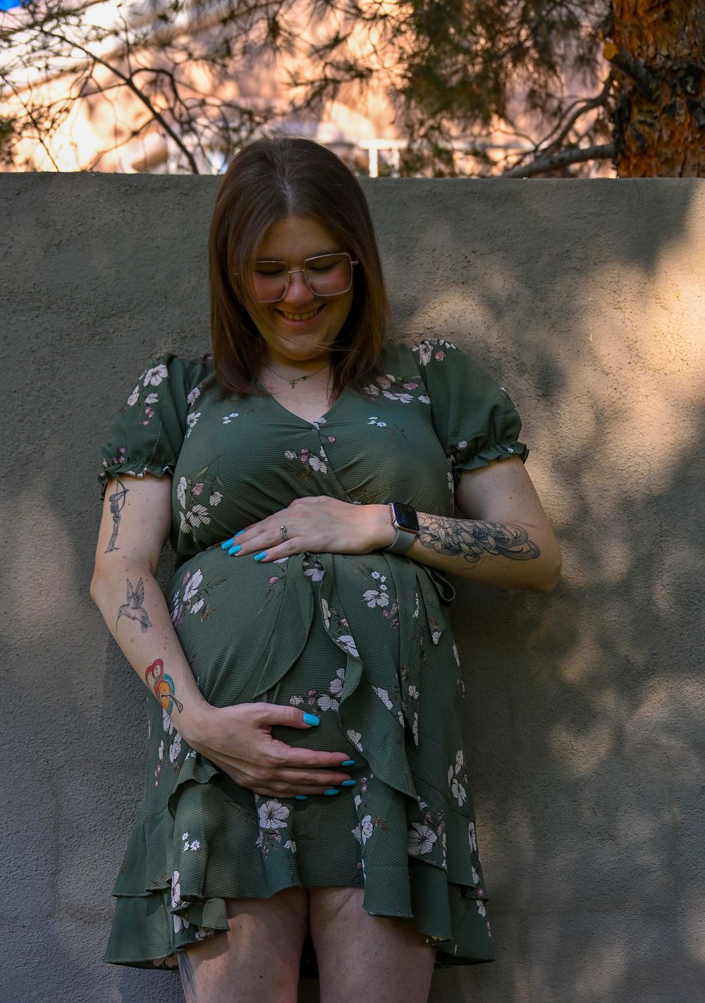 Smiling pregnant woman in green floral dress cradling baby bump against concrete wall outdoors.