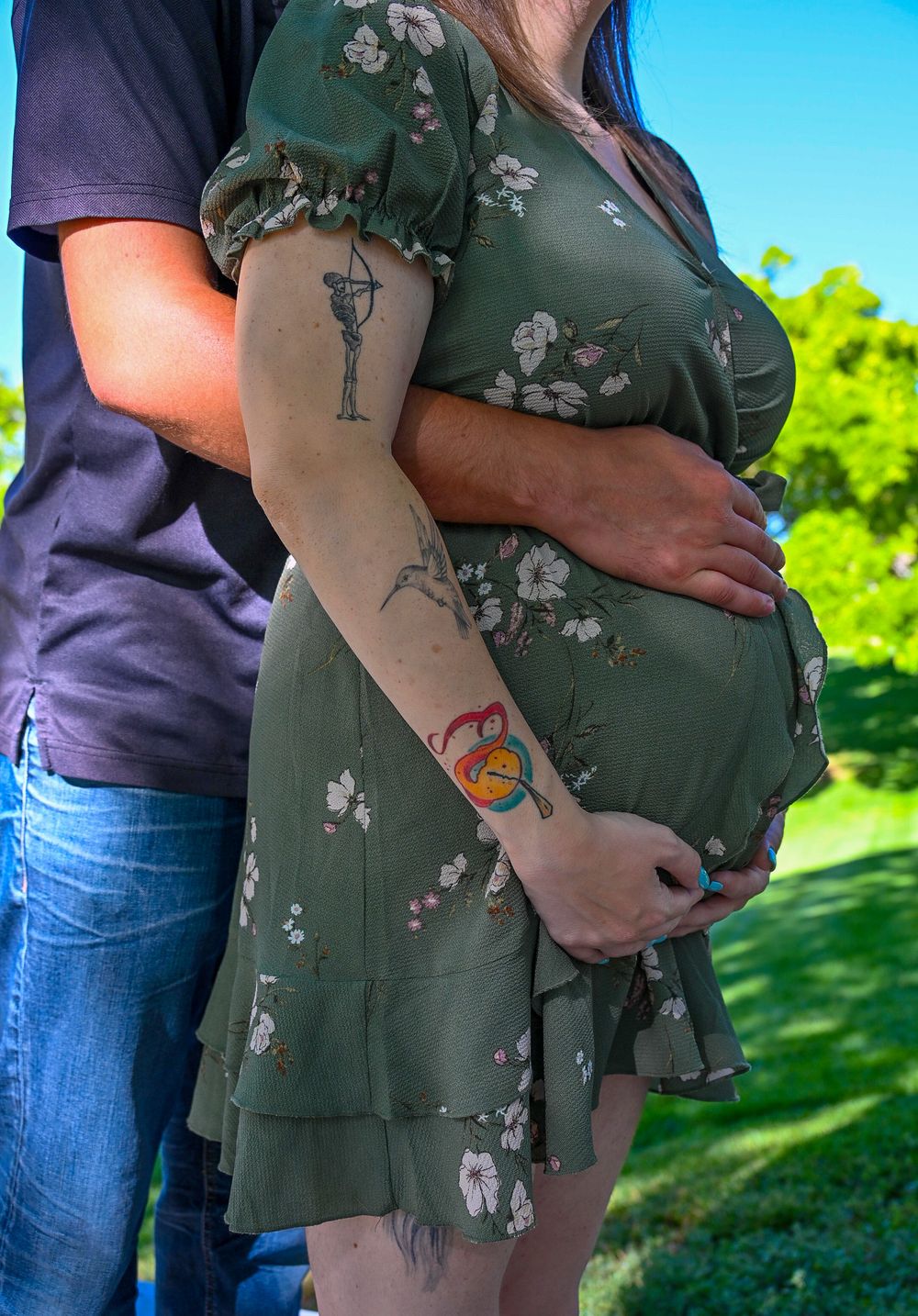 Pregnant woman in green floral dress with tattoos embraced by partner outdoors in sunny park.