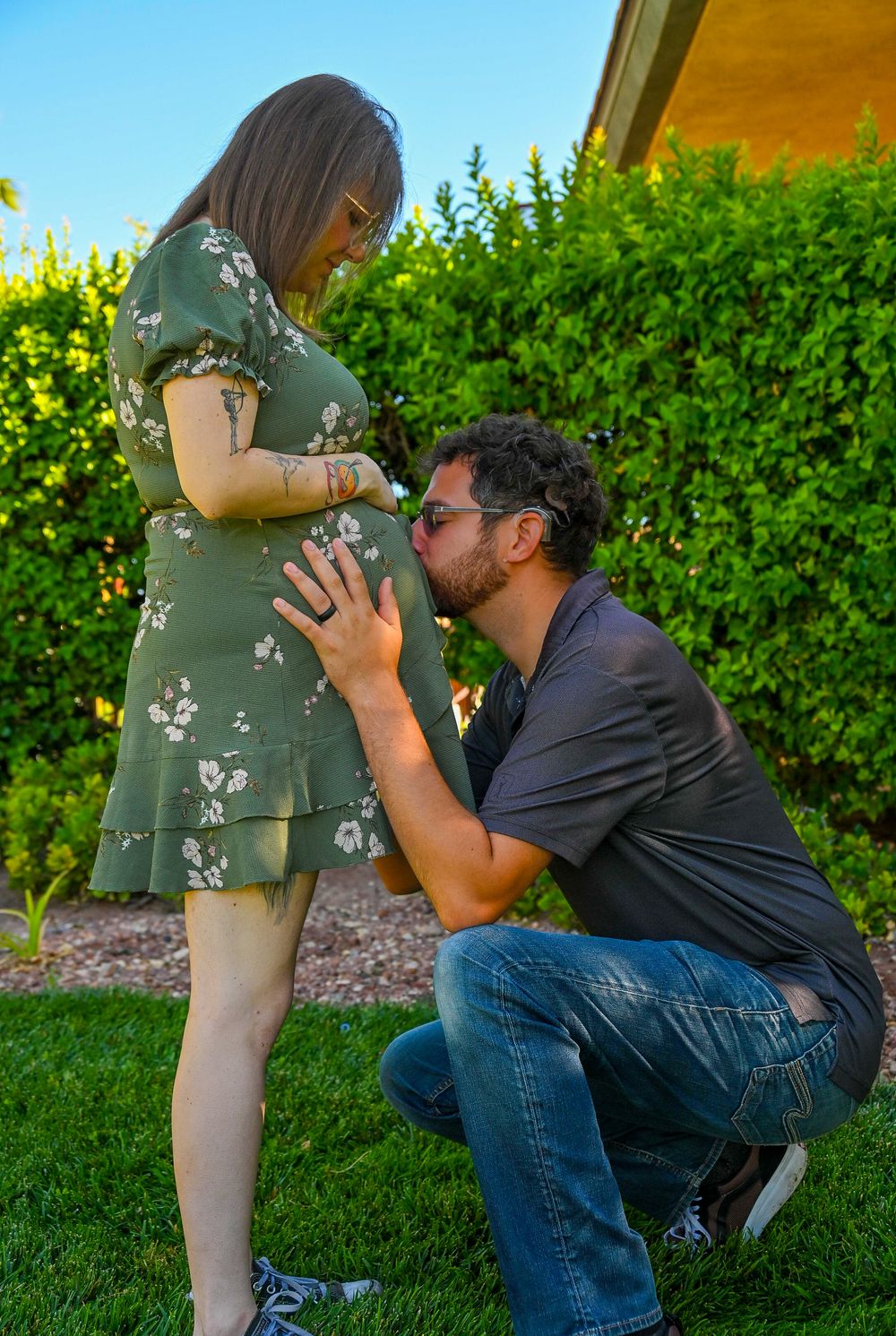 Man kneeling and kissing pregnant woman's belly outdoors in lush green garden during maternity photo session.