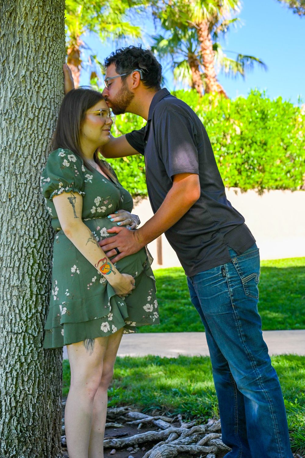Couple sharing tender kiss near tree trunk outdoors, man gently holding pregnant woman's baby bump.