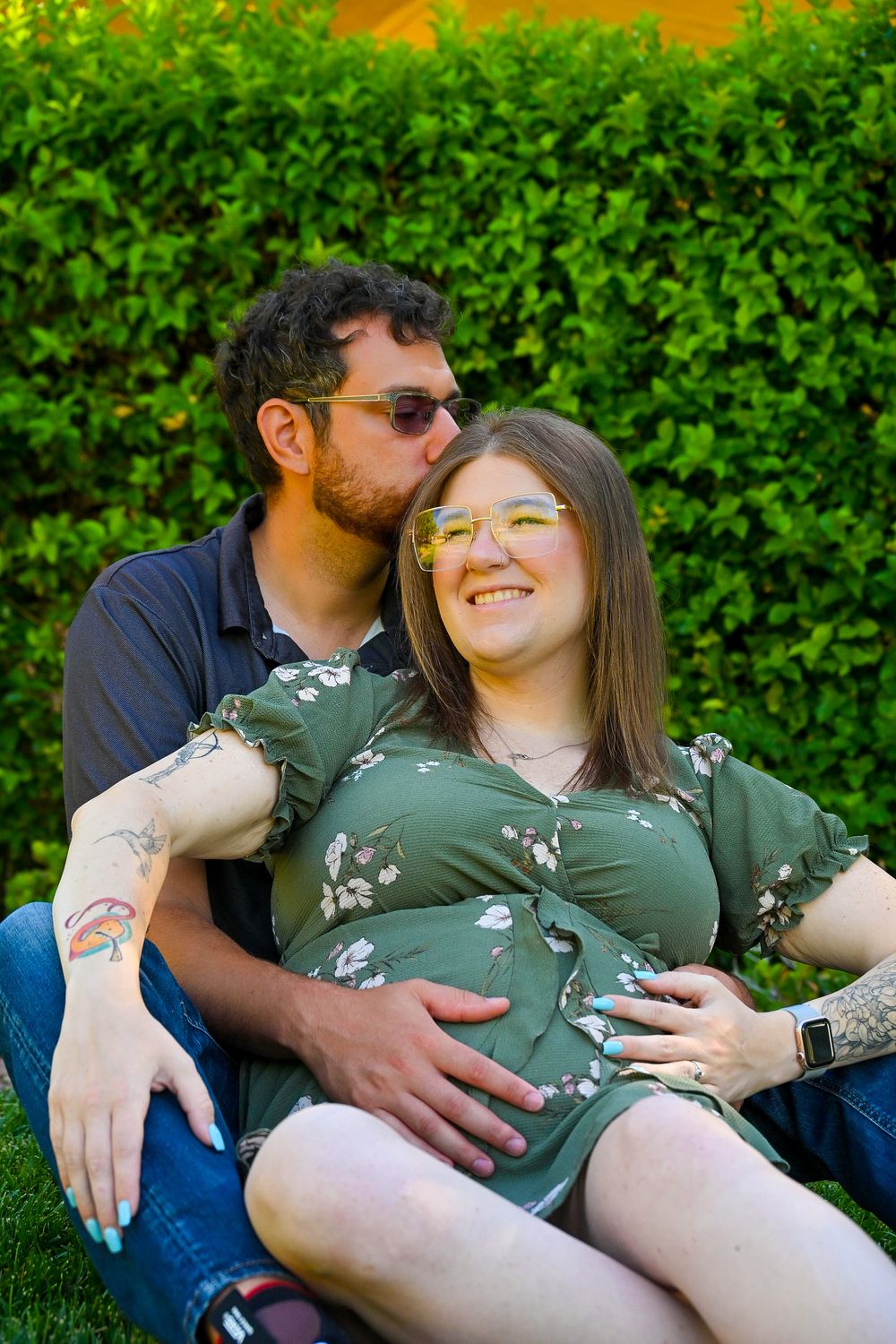 Happy pregnant woman in green floral dress smiling as partner kisses her cheek on grass.