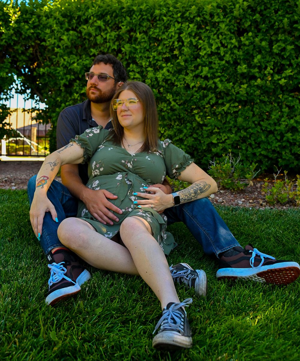 Couple sitting on grass together, both holding pregnant belly during outdoor maternity photo shoot.