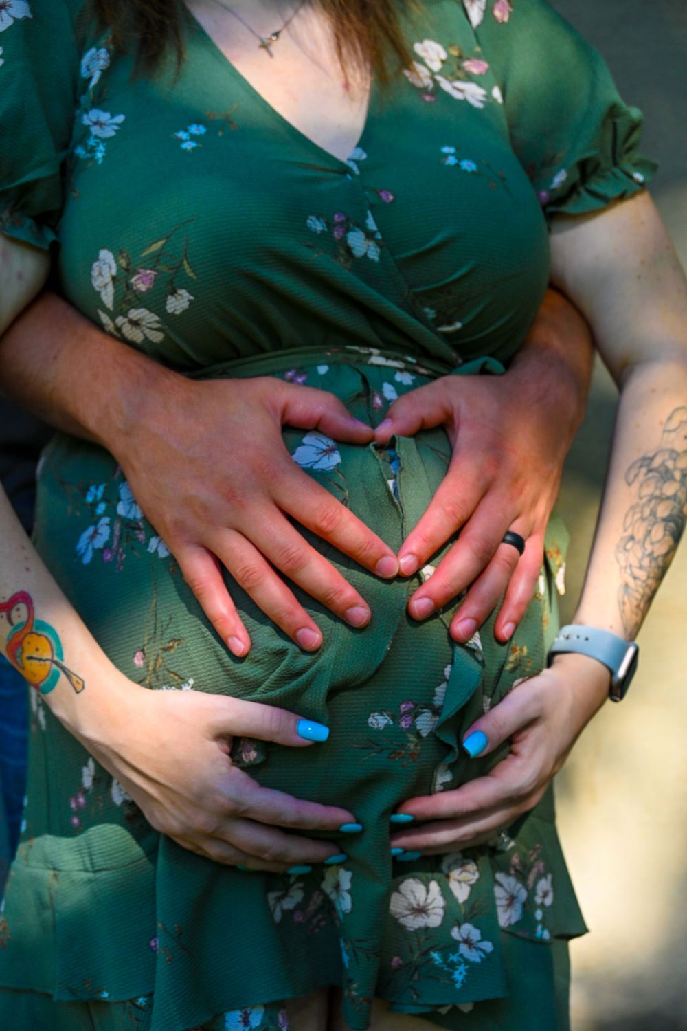Couple forming heart shape with hands on pregnant belly, woman wearing green floral dress with blue nails.