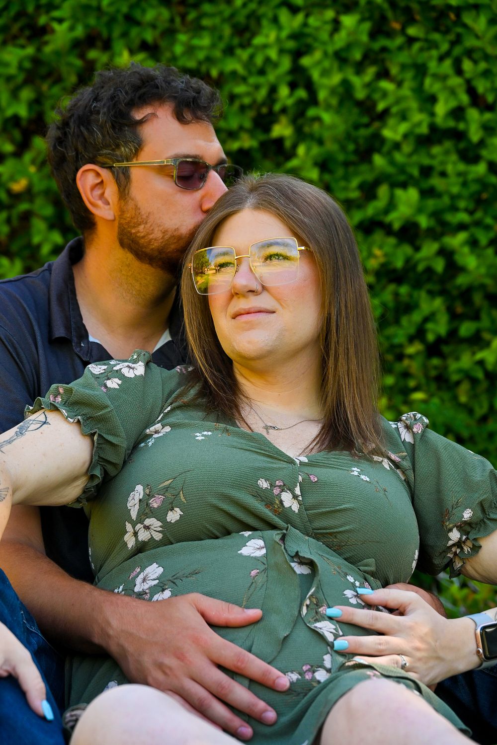 Man kissing pregnant partner on cheek as she smiles, both sitting against green hedge backdrop.
