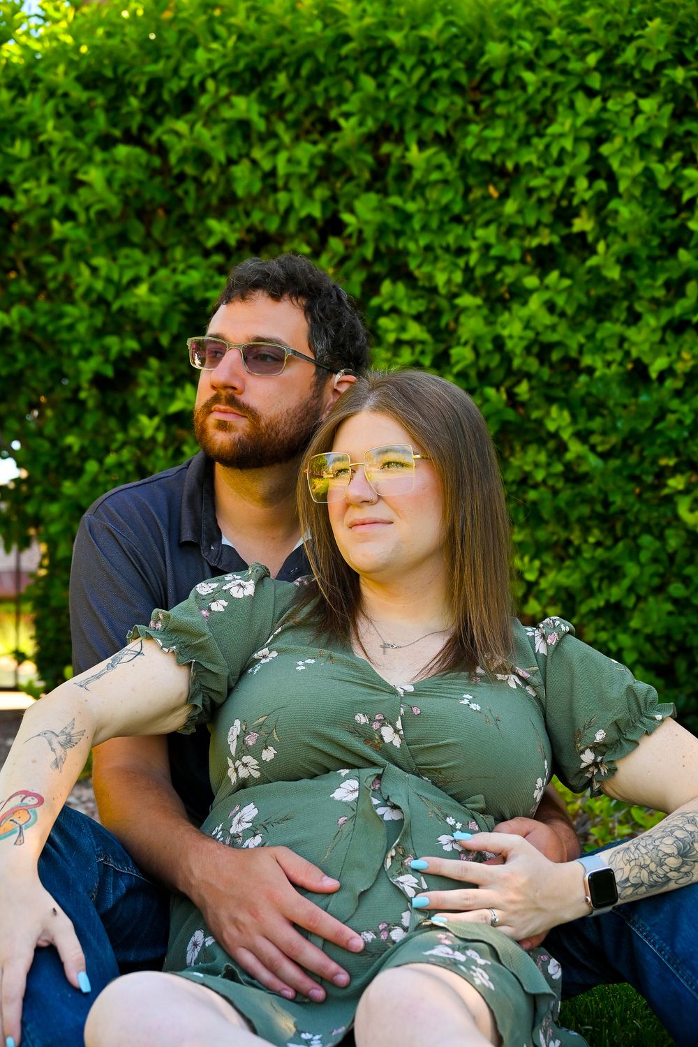 Couple sitting together against green hedge, man embracing pregnant woman during outdoor maternity session.