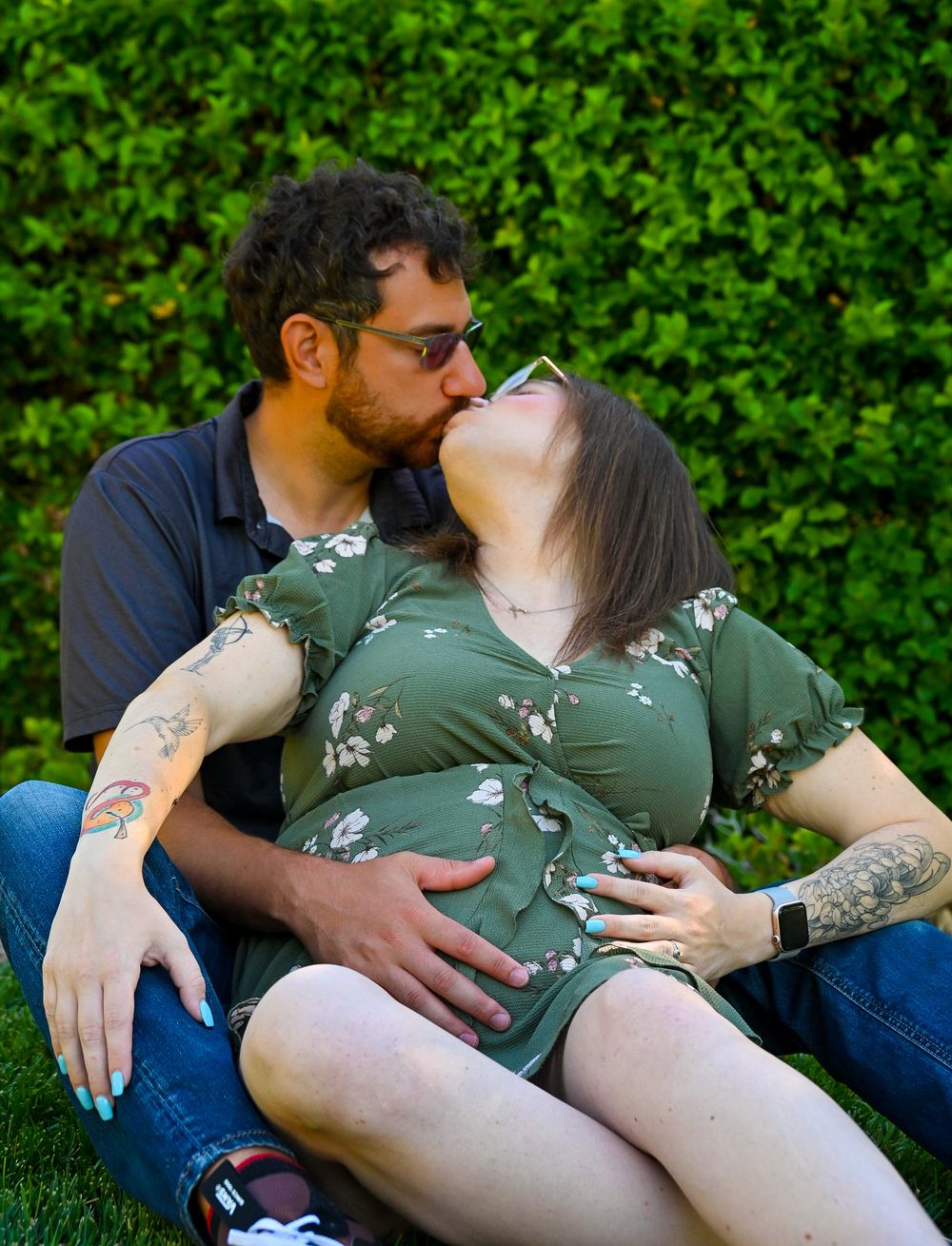 Couple sharing romantic kiss while sitting on grass during outdoor pregnancy photo session.