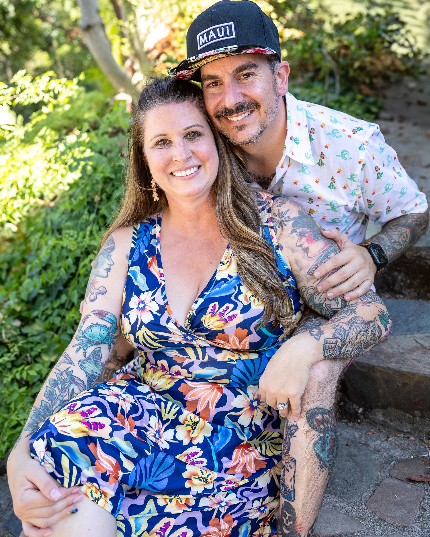 A couple in coordinating floral outfits pose together outdoors, with the person wearing a cap and tattoos on their arms.