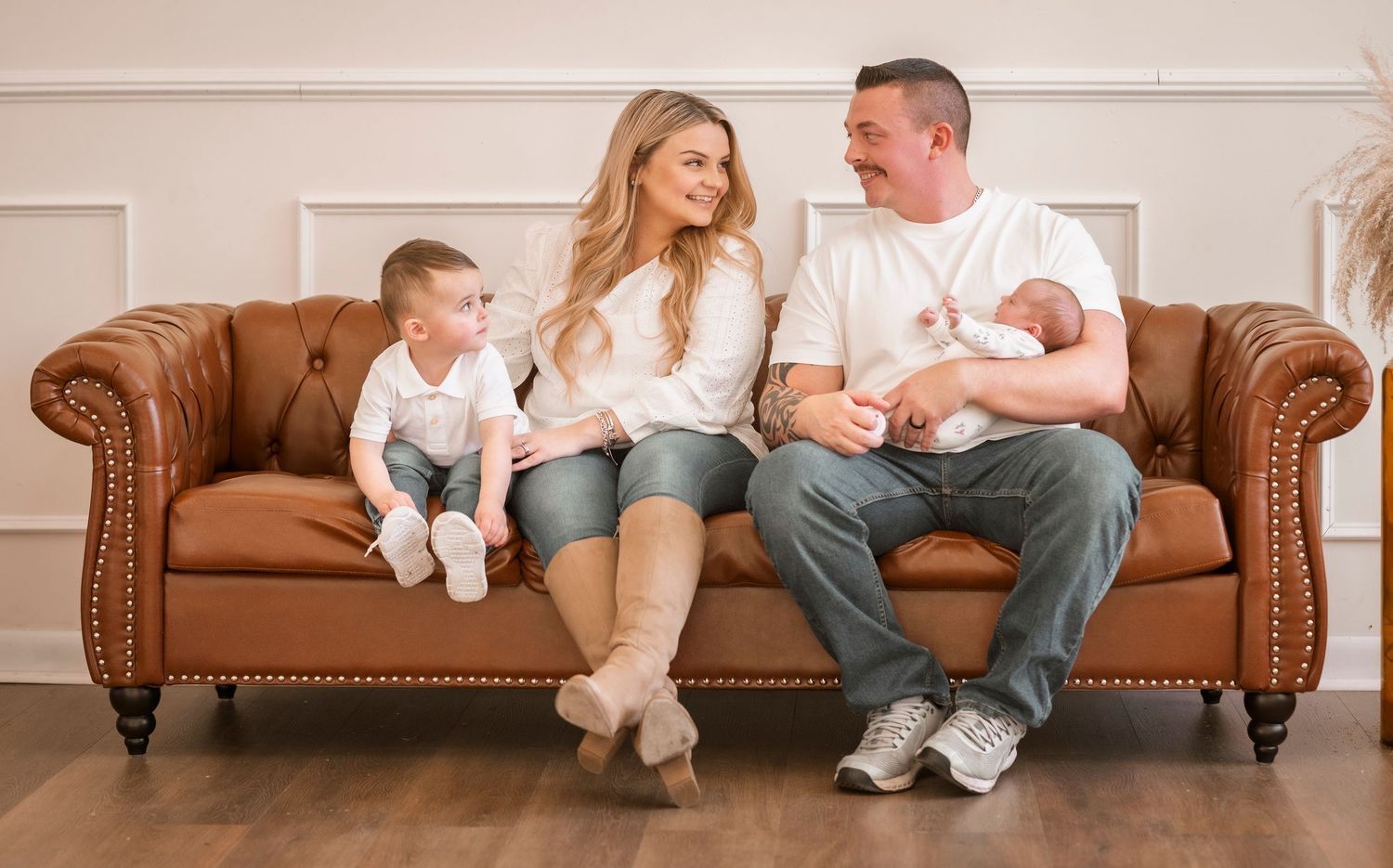 A family sits together on a brown leather couch in white and neutral colored clothes for a casual photo session.