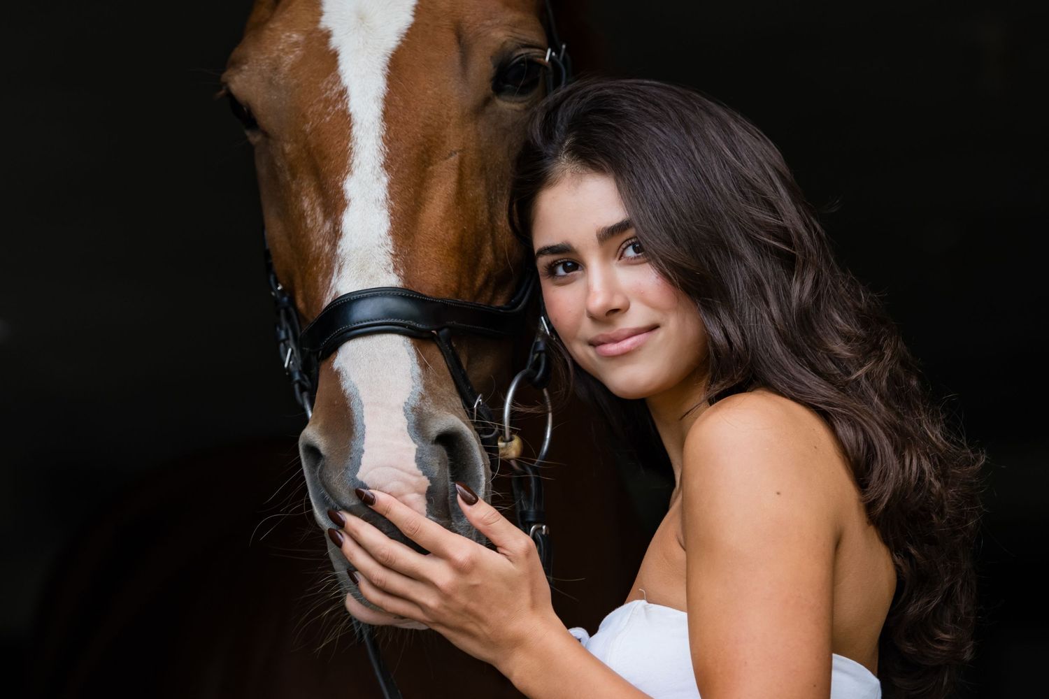 A brown and white horse stands with someone in a white top against a dark background.