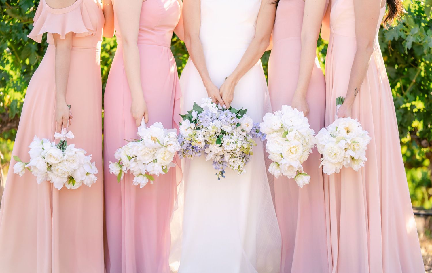 Row of pink bridesmaid dresses with white and purple bouquets against a sunny backdrop.