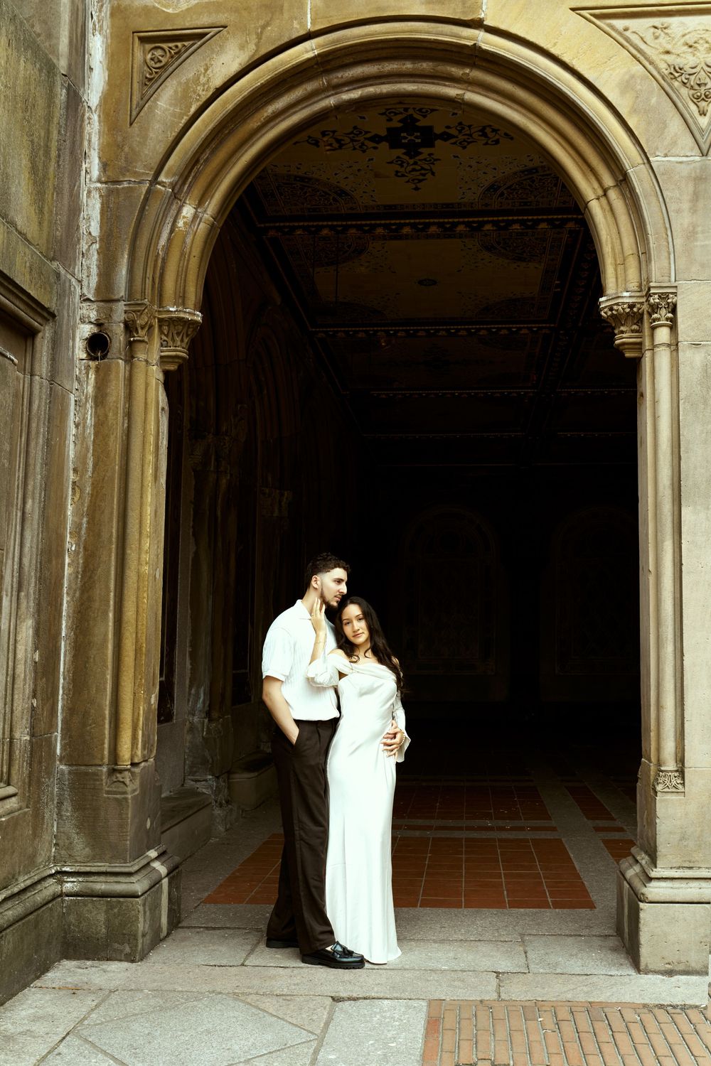Elegant couple in white poses beneath historic stone archway.