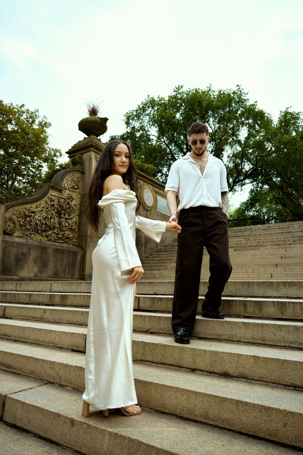 A couple in elegant white outfits poses on grand stone steps outdoors.