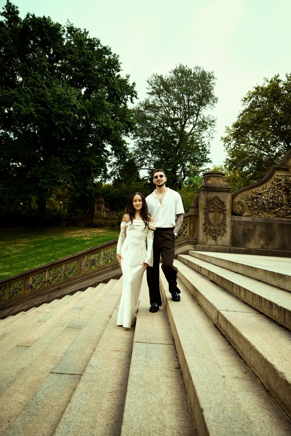 A couple in coordinated formal wear descends ornate stone steps in a park setting.