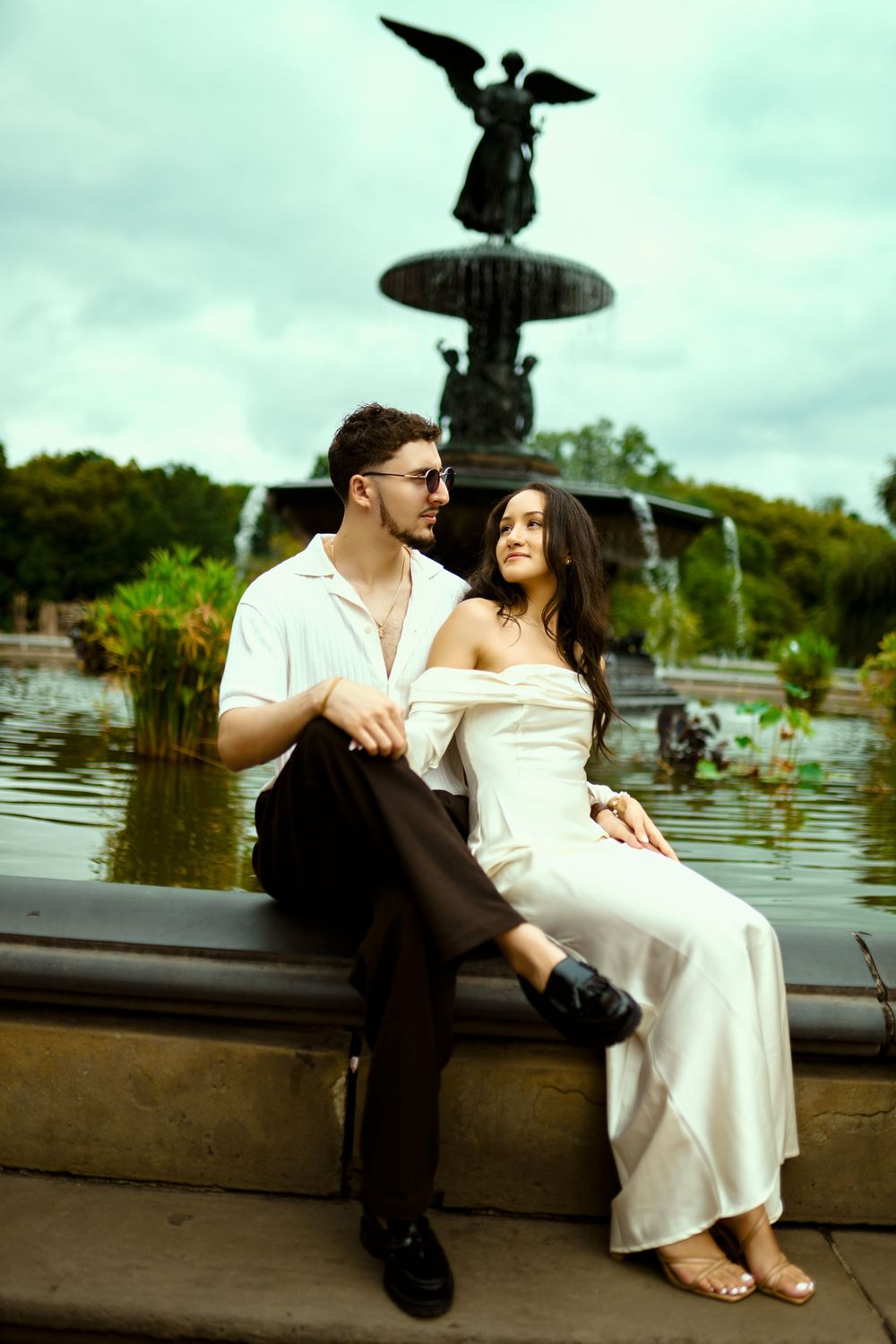 A couple sits by Bethesda Fountain in Central Park with the Angel statue visible in background.