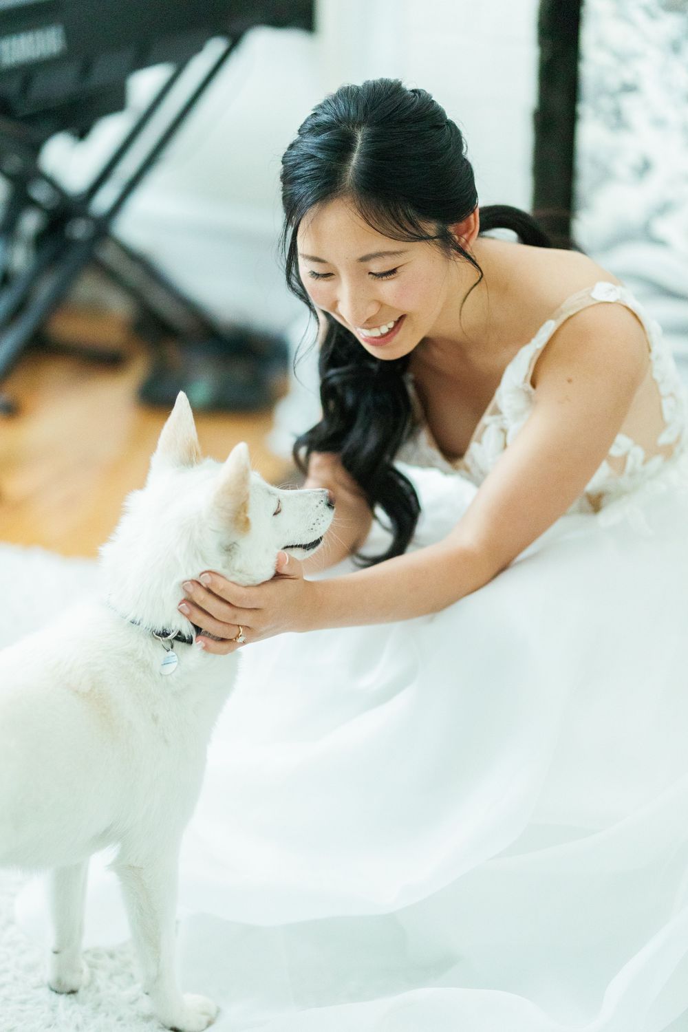 A bride in a flowing white gown bends down to pet her small white dog during a photoshoot.