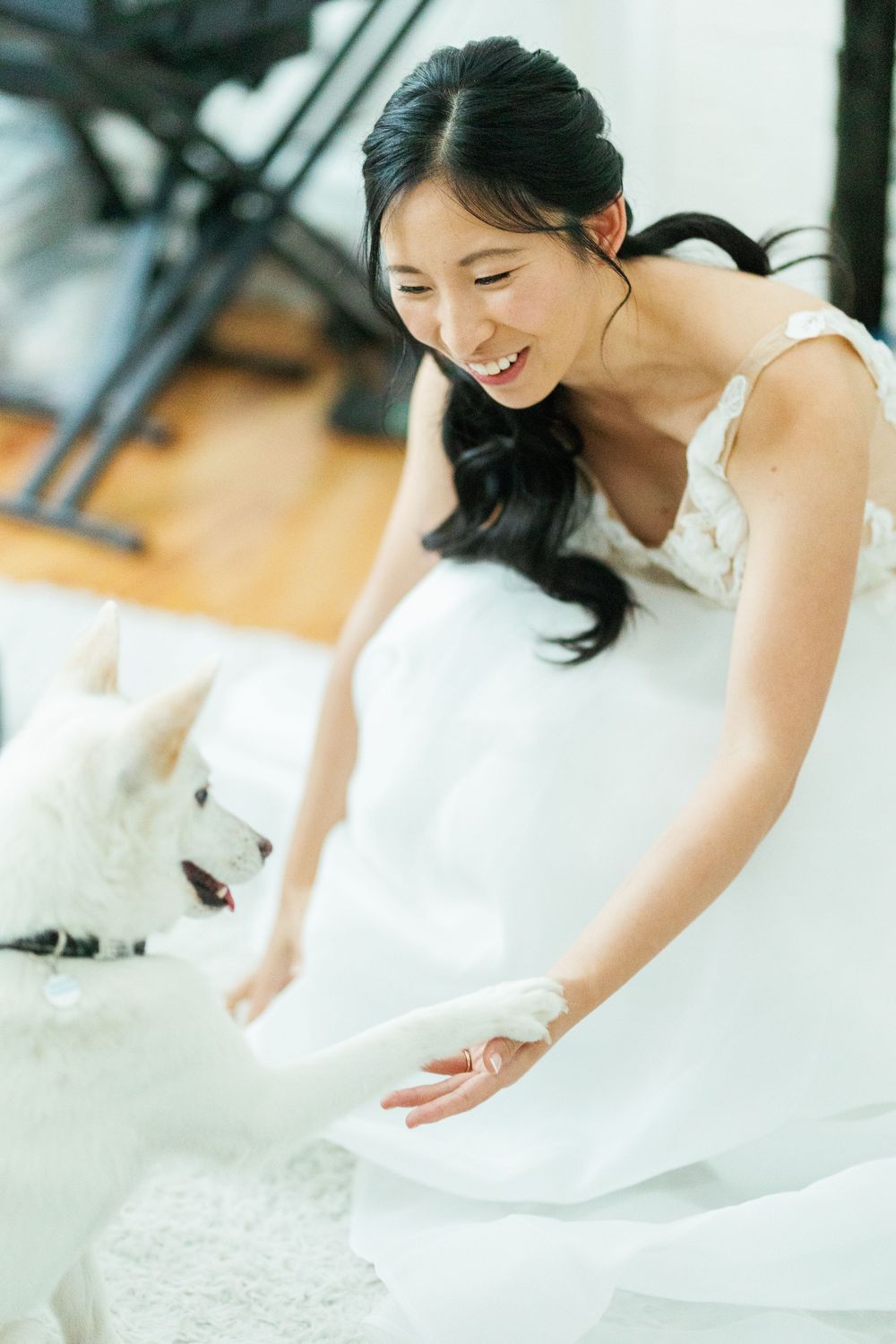 A bride in a white dress smiles joyfully while sitting on the floor with a small white dog.