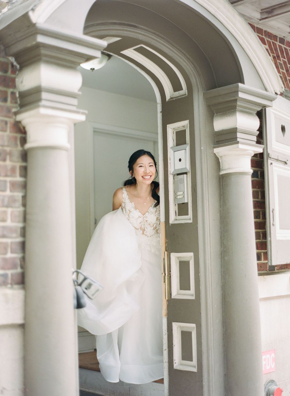 A person in a flowing white wedding dress spins joyfully between elegant white columns in a classic brick building.