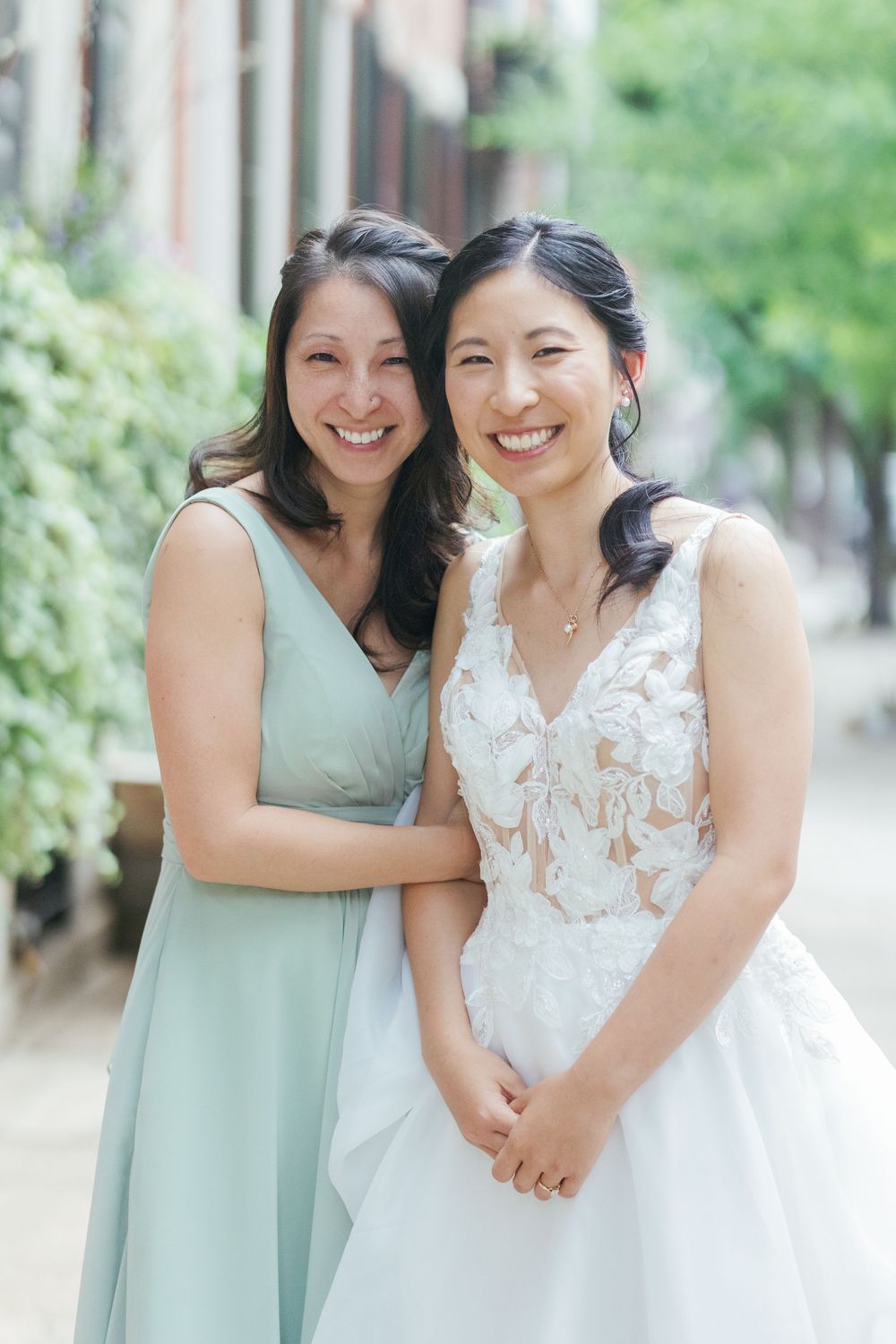 Two women embrace and smile joyfully in coordinating mint green bridesmaid dress and white lace wedding gown outdoors.