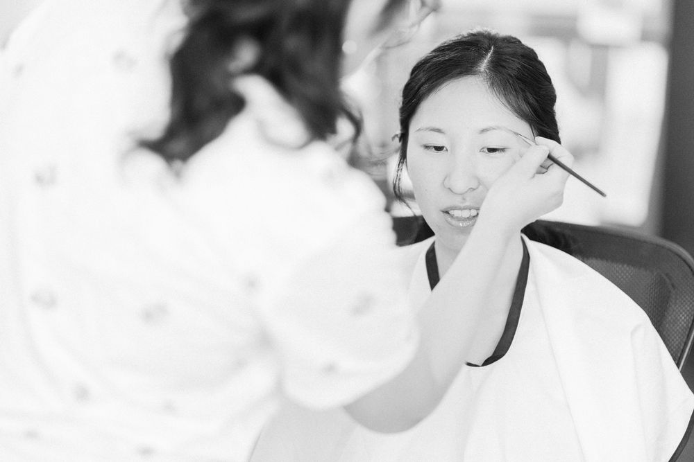 Black and white photo of person having makeup applied by makeup artist in bright setting.