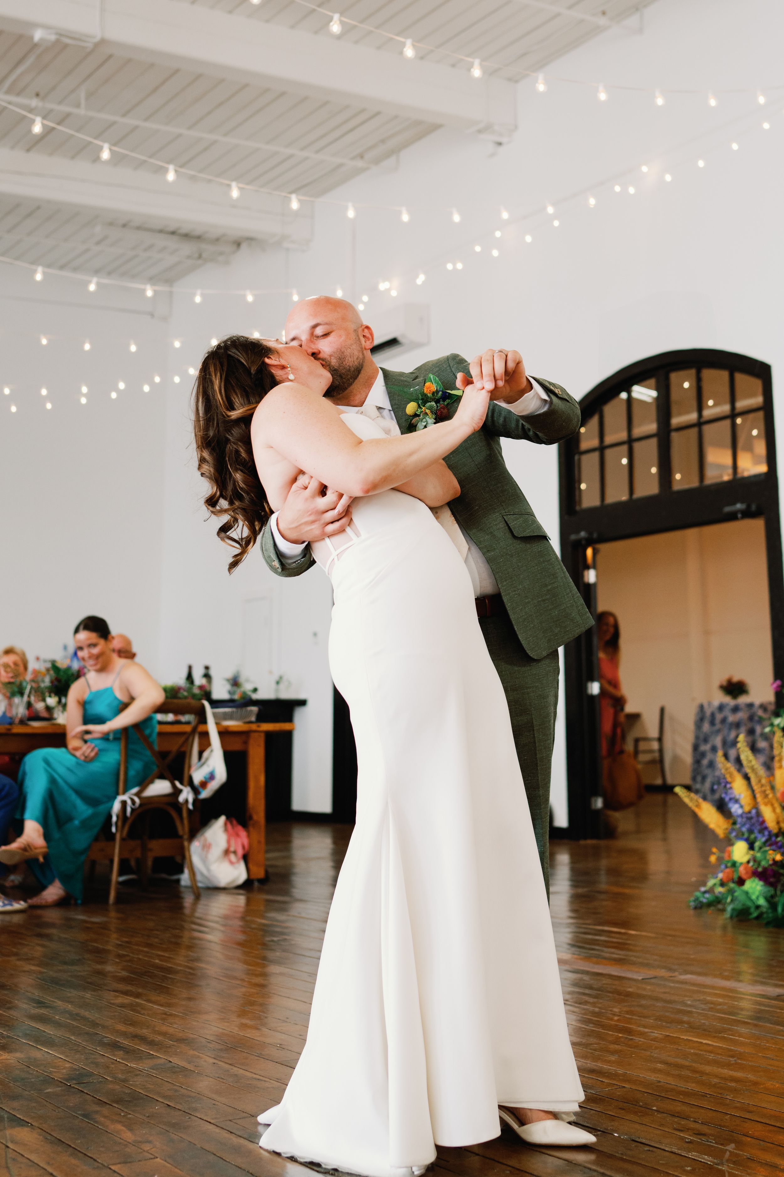 A couple dances together in an industrial style venue with string lights and wooden floors during a wedding celebration.