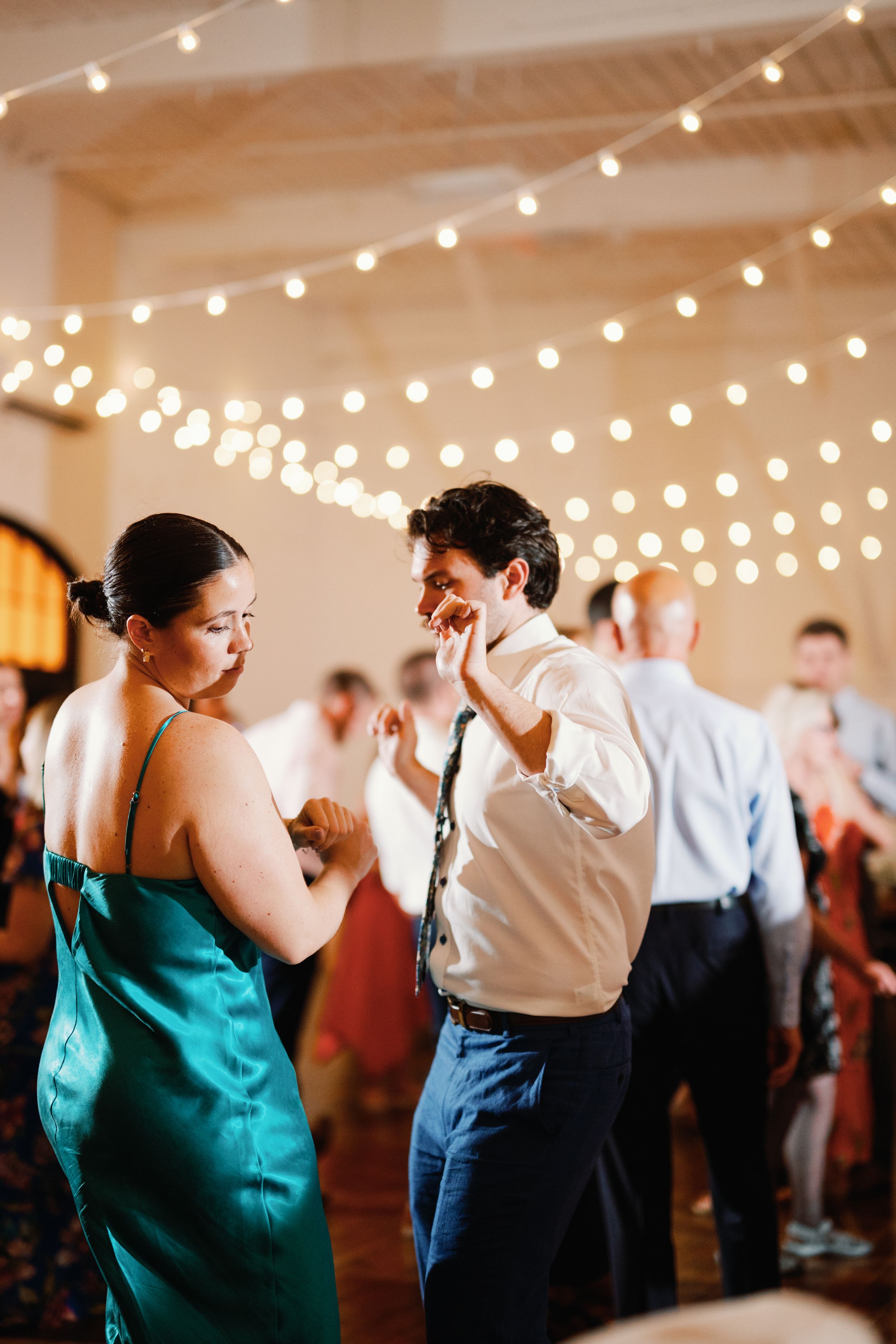 Two people dance together at a wedding reception under warm string lights in an indoor venue.