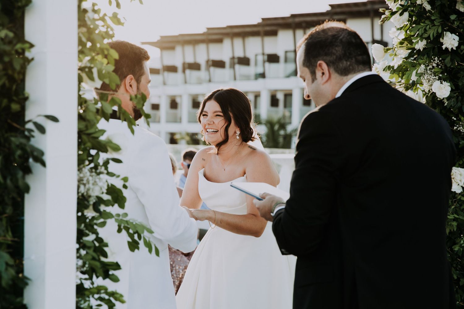 A romantic outdoor wedding ceremony with guests seated in white chairs facing a floral arch overlooking the ocean.