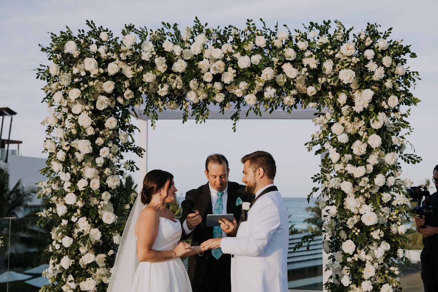 Wedding party celebrating under an elegant white floral arch during a seaside ceremony.