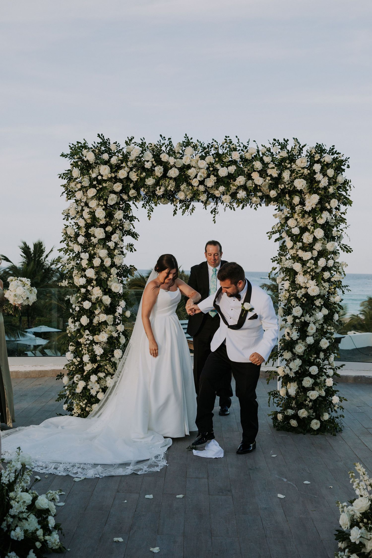 Wedding party members playfully posing and dancing under a luxurious white floral arch installation.