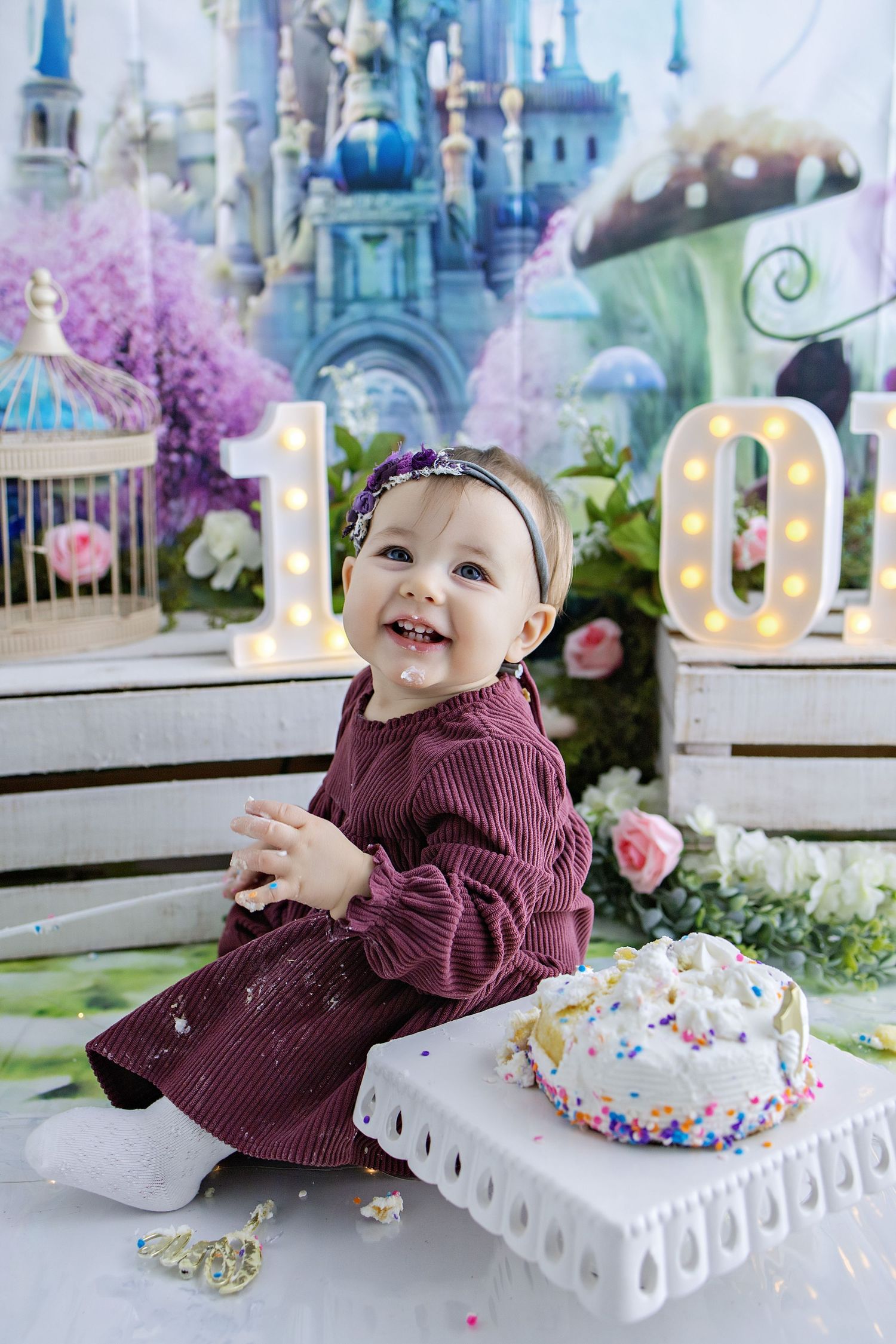 A baby in a blue dress sits on lace fabric outdoors enjoying a white birthday cake against a sunset bokeh background.