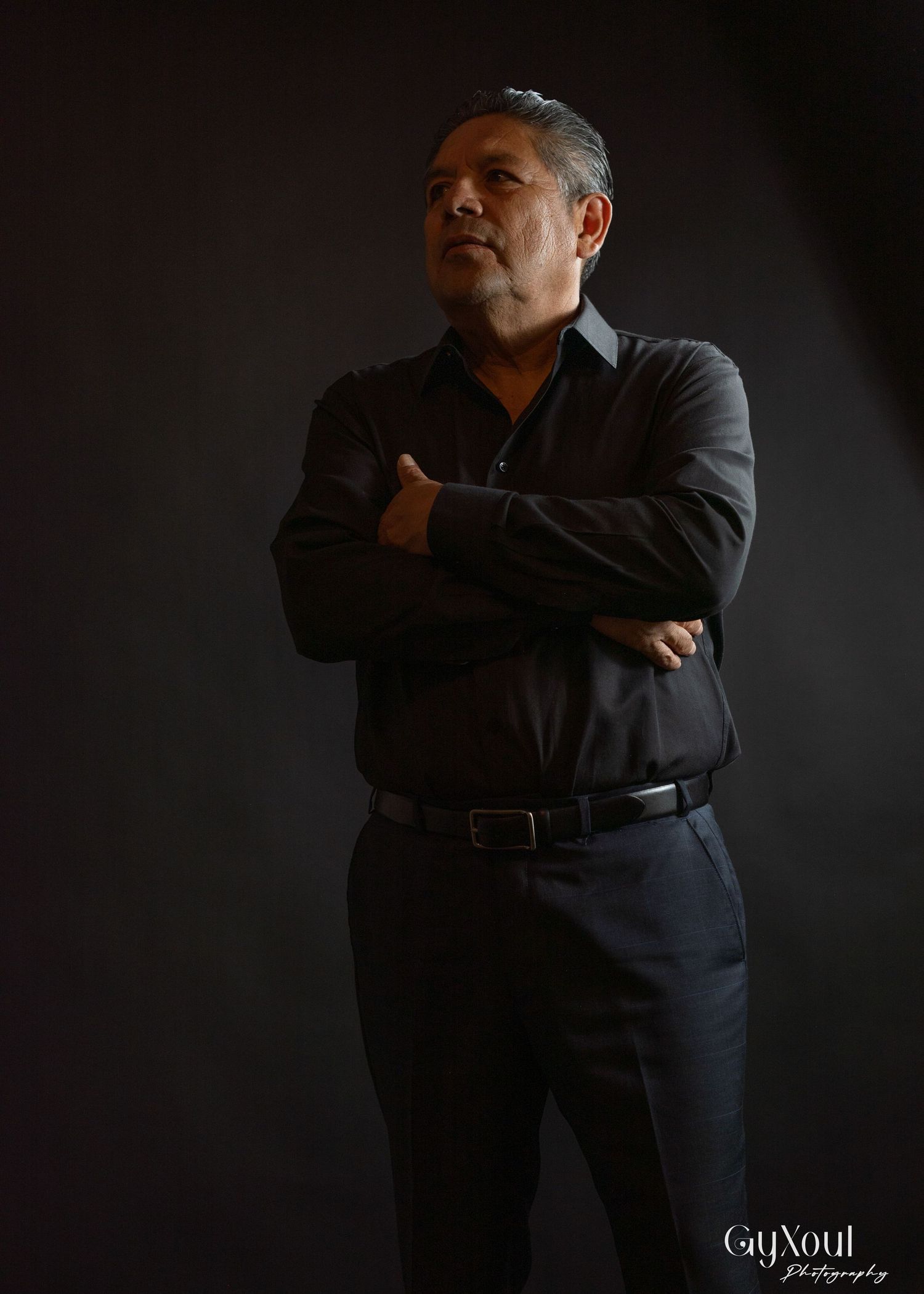 A person in dark business attire stands with arms crossed in a dramatic low-key portrait against a black background.