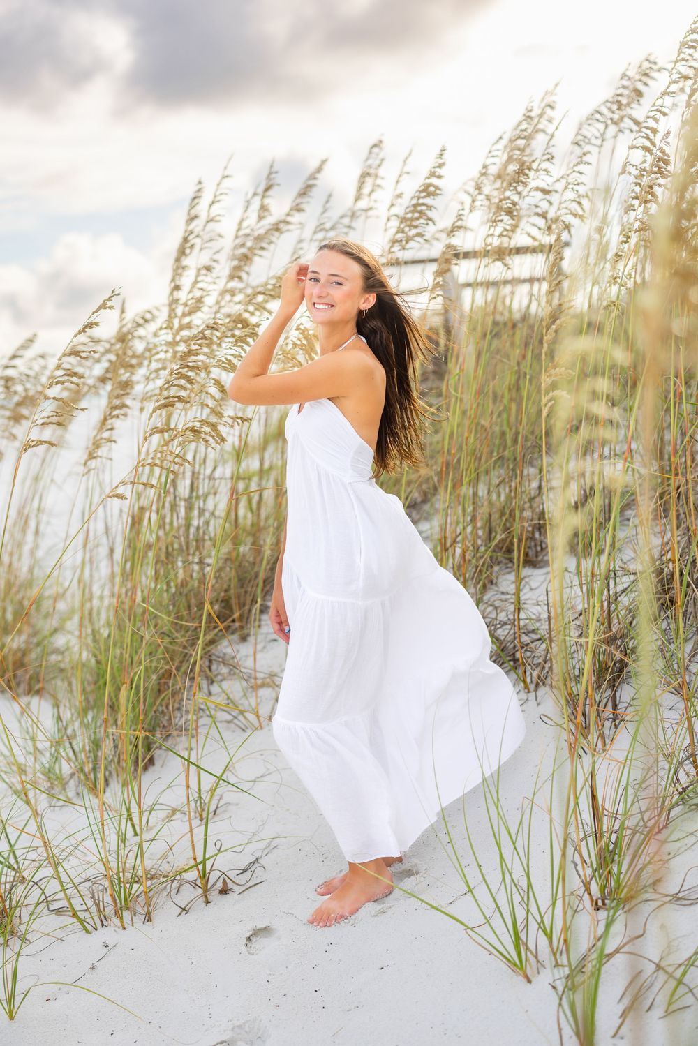 A woman in a flowing white dress poses gracefully among sea oats on a sandy beach with cloudy skies above.