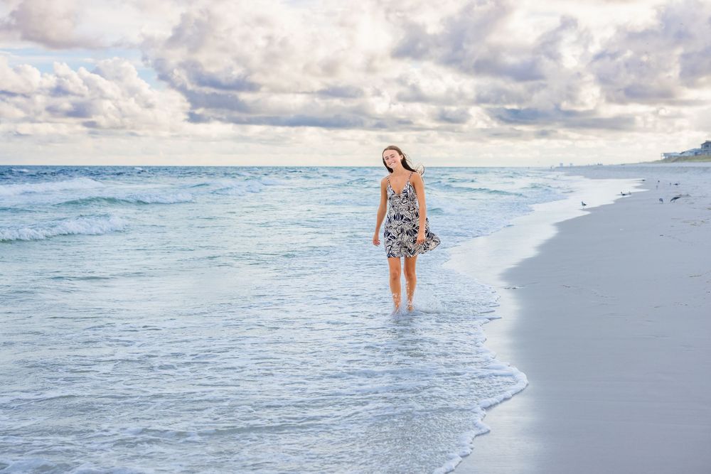 A person walks along the shoreline of a beautiful beach at sunset with waves rolling in and cloudy skies overhead.