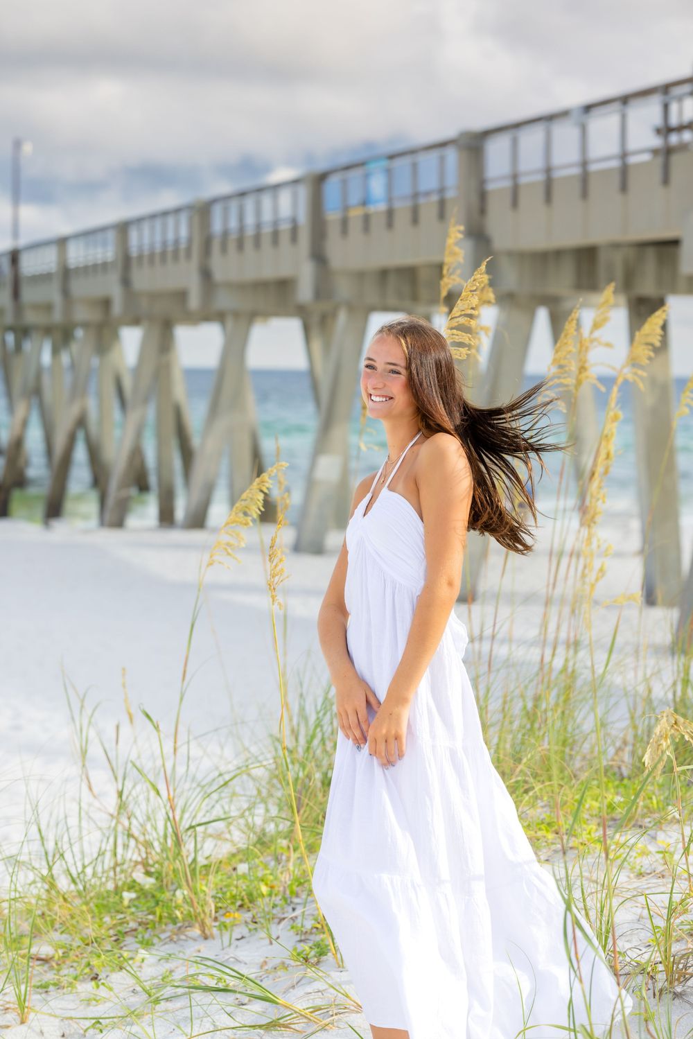 A woman in a white sundress smiles by a pier surrounded by beach grass and white sand.