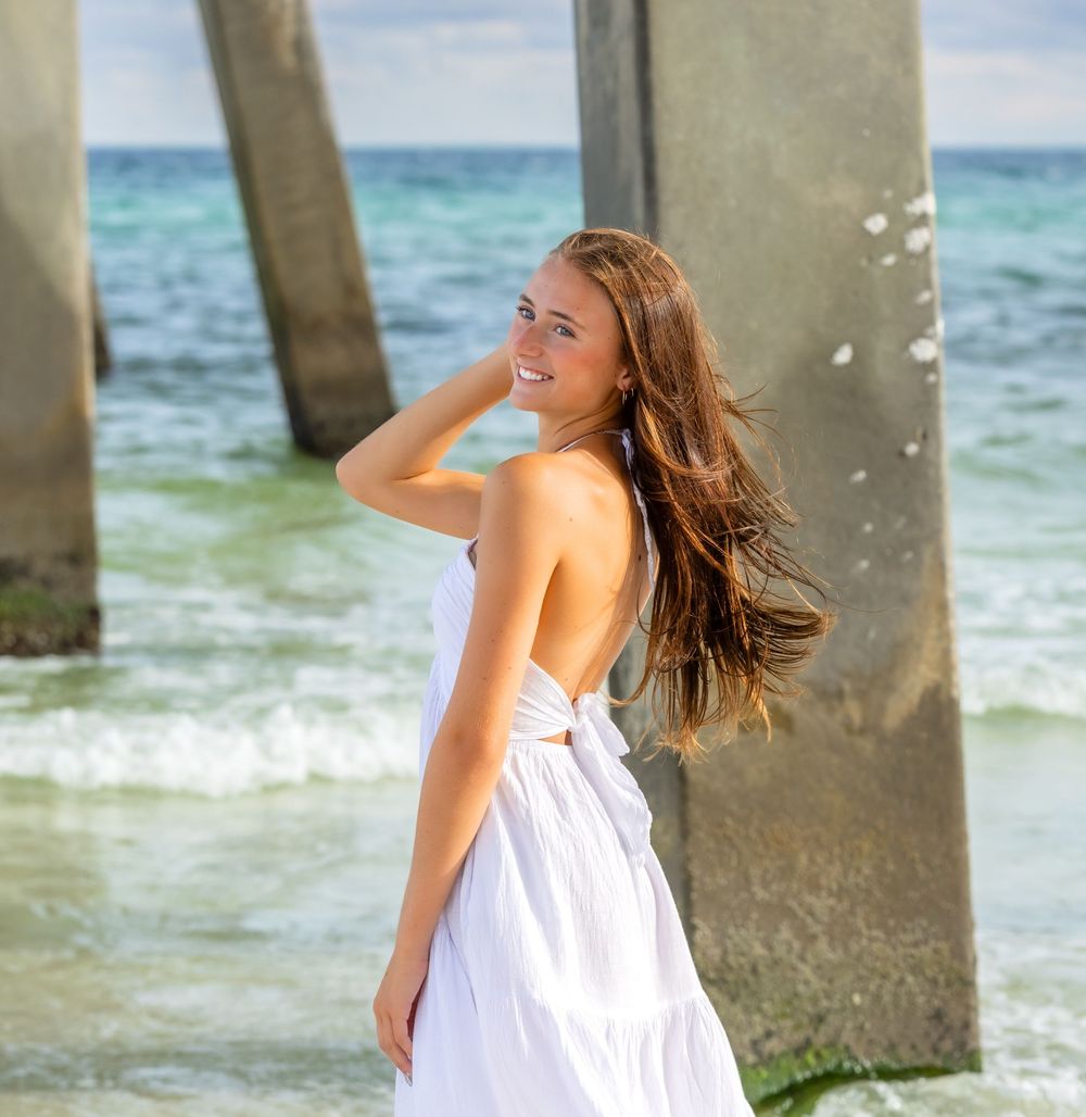 A person in a white sundress poses near a pier on a tropical beach with turquoise ocean waters in the background.