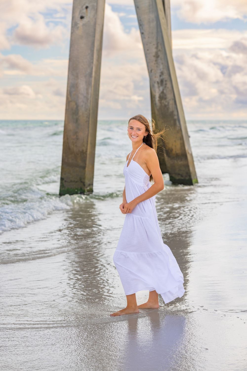 A flowing white dress catches the sea breeze as someone poses near wooden pier pillars at the beach.