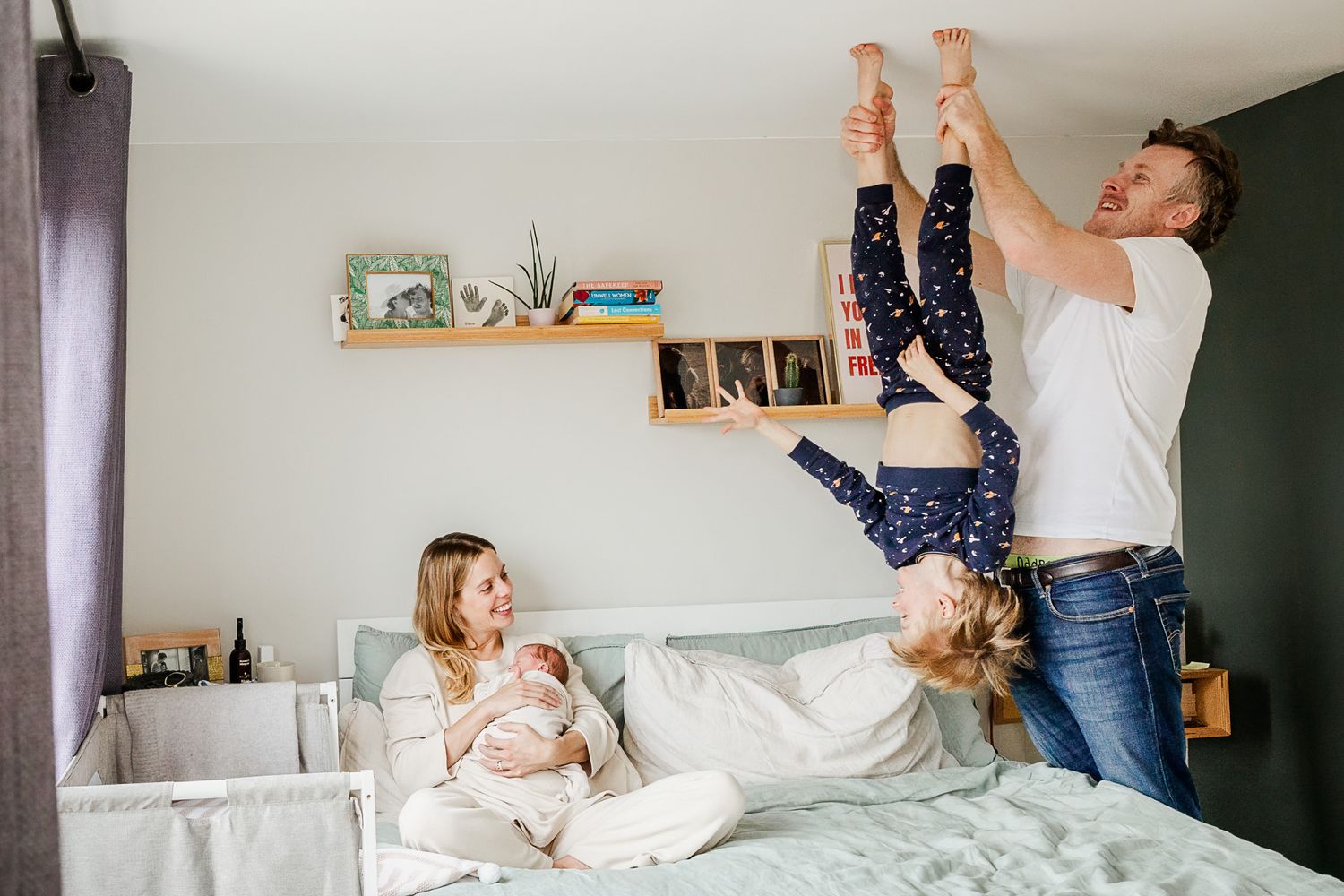 A dad plays and holds his son upside down and smiles as he relaxes during a Bristol newborn photoshoot at home. Mum cuddles their new baby.