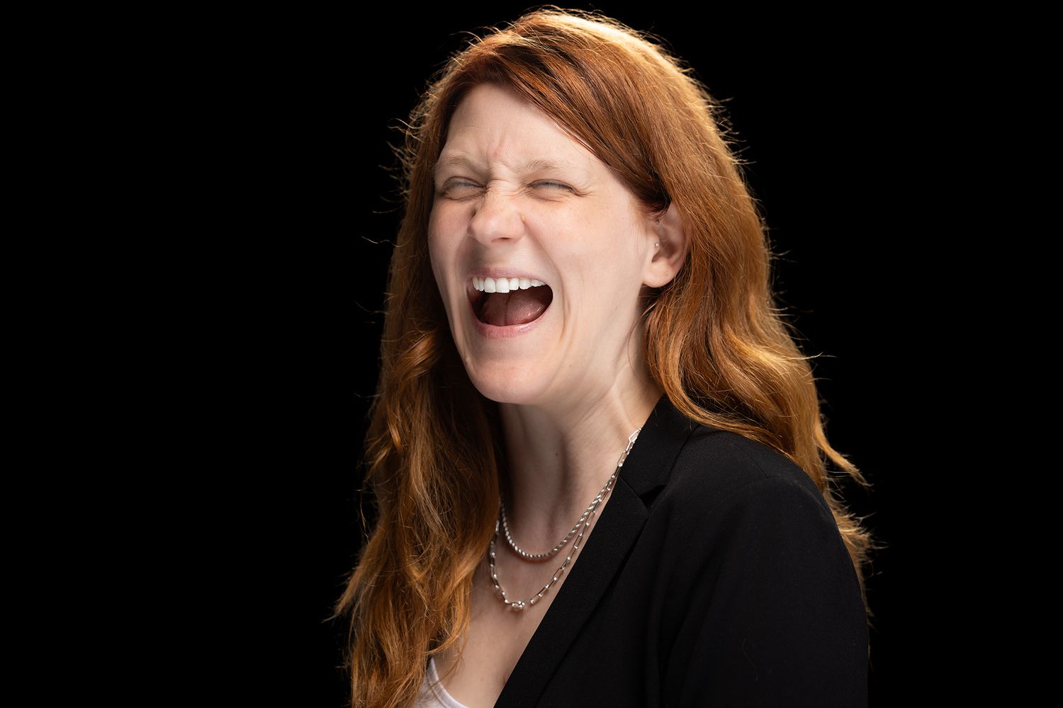 A woman with long red hair in a black blazer laughs joyfully against a dark background.